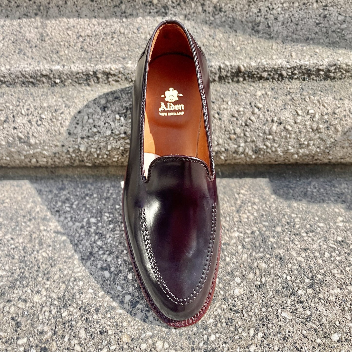A polished dark leather loafer crafted from Color 8 Shell Cordovan, this Alden Madison D1118 - Vanderbilt Slip On boasts a sleek design with a branded insole that reads Alden New England, resting on a textured stone step and casting a soft shadow in the sunlight.