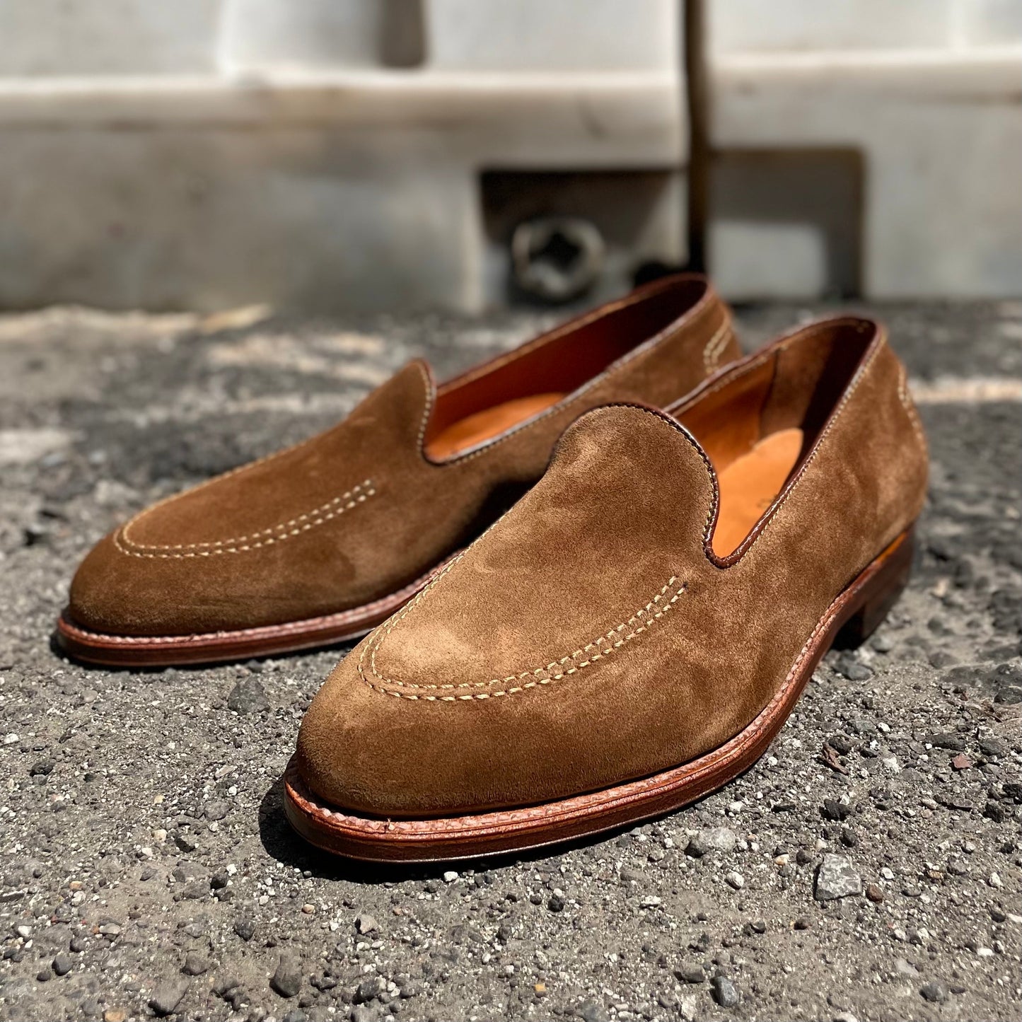 The Alden Madison D2101 - Vanderbilt Slip On in snuff suede features stitched detailing and a leather sole, resting on a textured surface with an elegant blurred white structure in the background.