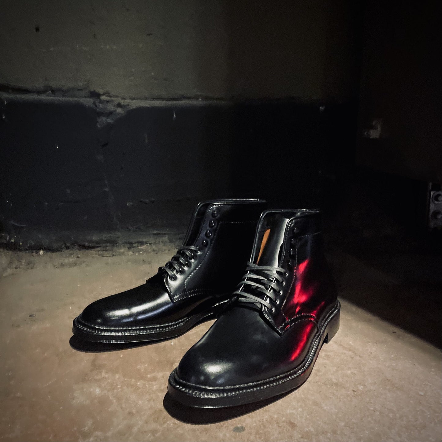 The D6842H Director Boot in Black Shell Cordovan from Alden Madison rests on a dimly lit concrete floor, casting shadows against a dark wall.