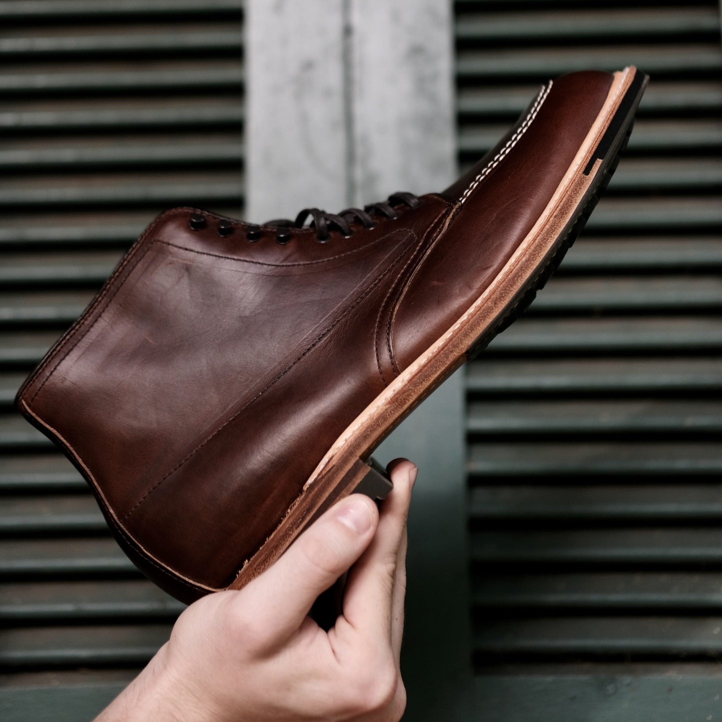 A hand holds an Alden Madison 403C - Indy Boot in brown Chromexcel leather, featuring detailed stitching and a commando sole. The backdrop consists of horizontal metal slats.