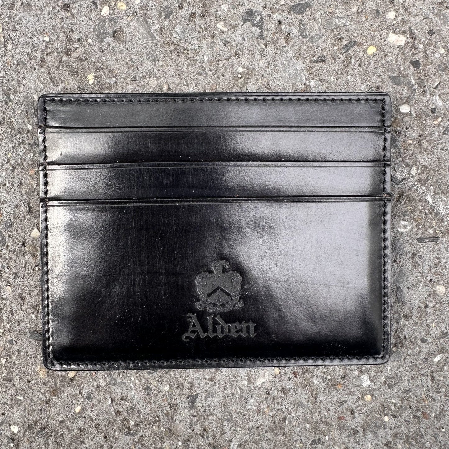 The Alden Madison LG 6009 is a black leather Shell Cordovan cardholder embossed with Alden and a crown logo, featuring six pockets for essentials, elegantly displayed on textured stone.