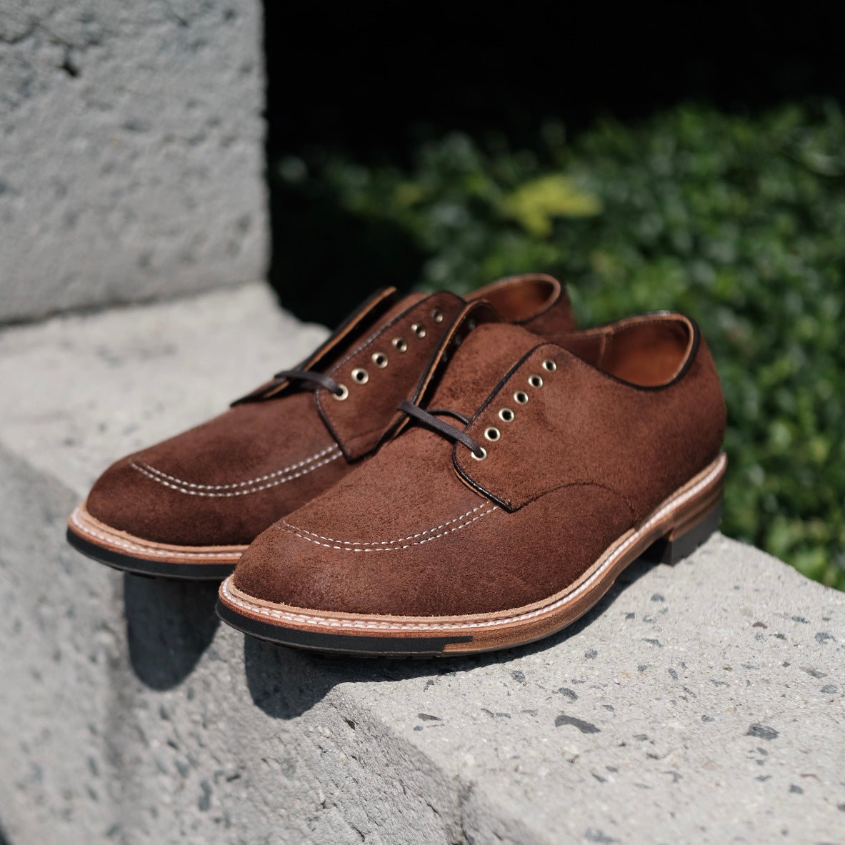 The Alden Madison D2604C - Indy Shoe in Tobacco Reverse Chamois, crafted in brown suede with the Trubalance Last, features eyelets, thick soles, dark laces, and contrasting light stitching on a concrete surface. Blurred green foliage provides the background.