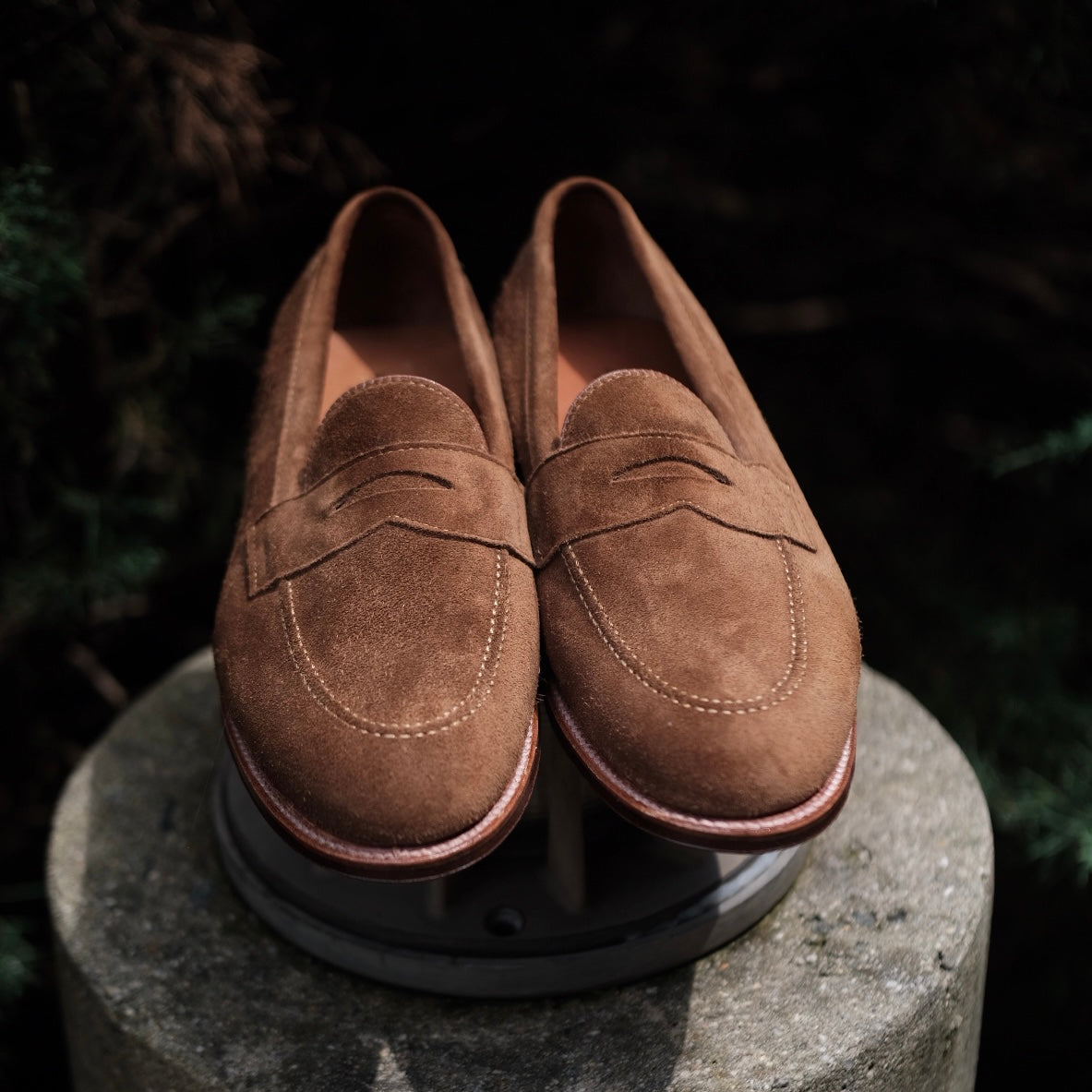 A pair of Alden Madisons 96941F - Flex Penny Loafers in Snuff Suede, featuring decorative stitching, elegantly perched on a concrete pedestal outdoors against a dark, leafy backdrop.
