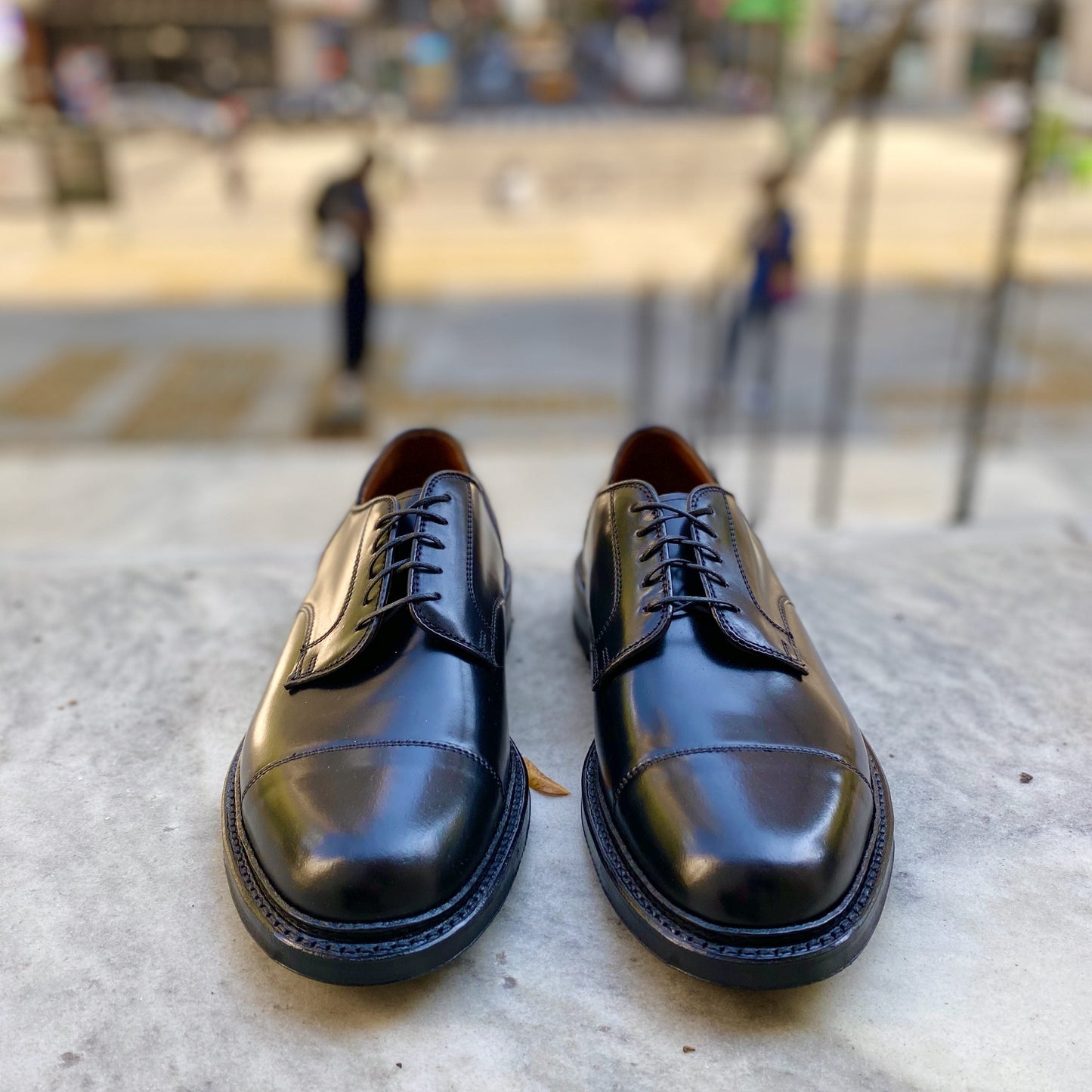 A pair of Alden Madisons 2175C - DeRosa Captoe Blucher in Black Shell, featuring a sleek design and Commando Sole, is set on a stone ledge. The blurred urban street scene background shows two people walking.