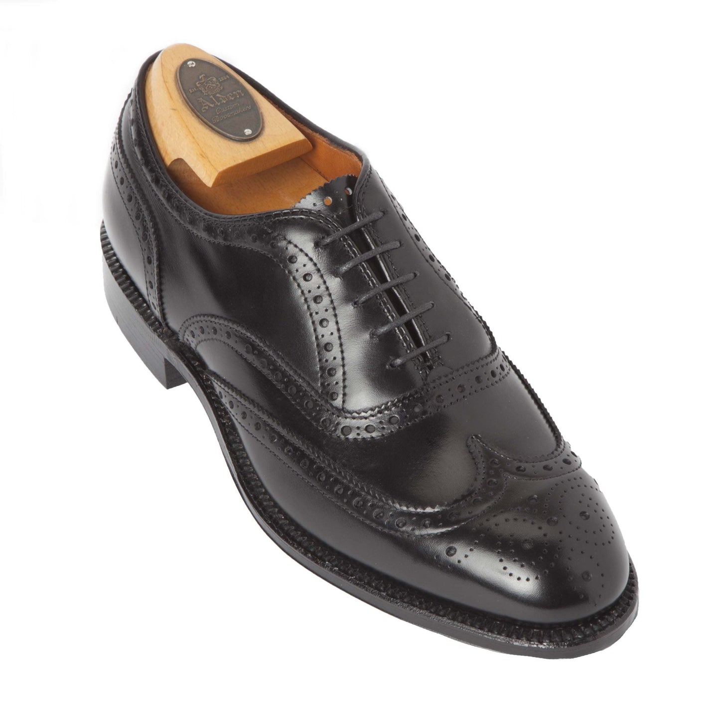 Alden Madisons 903 - Wing Tip in Bal Black Calfskin is showcased at an angle, featuring premium black calfskin, decorative perforations, and a wingtip design. It has black laces and includes a wooden shoe tree, all set on the refined Hampton Last.