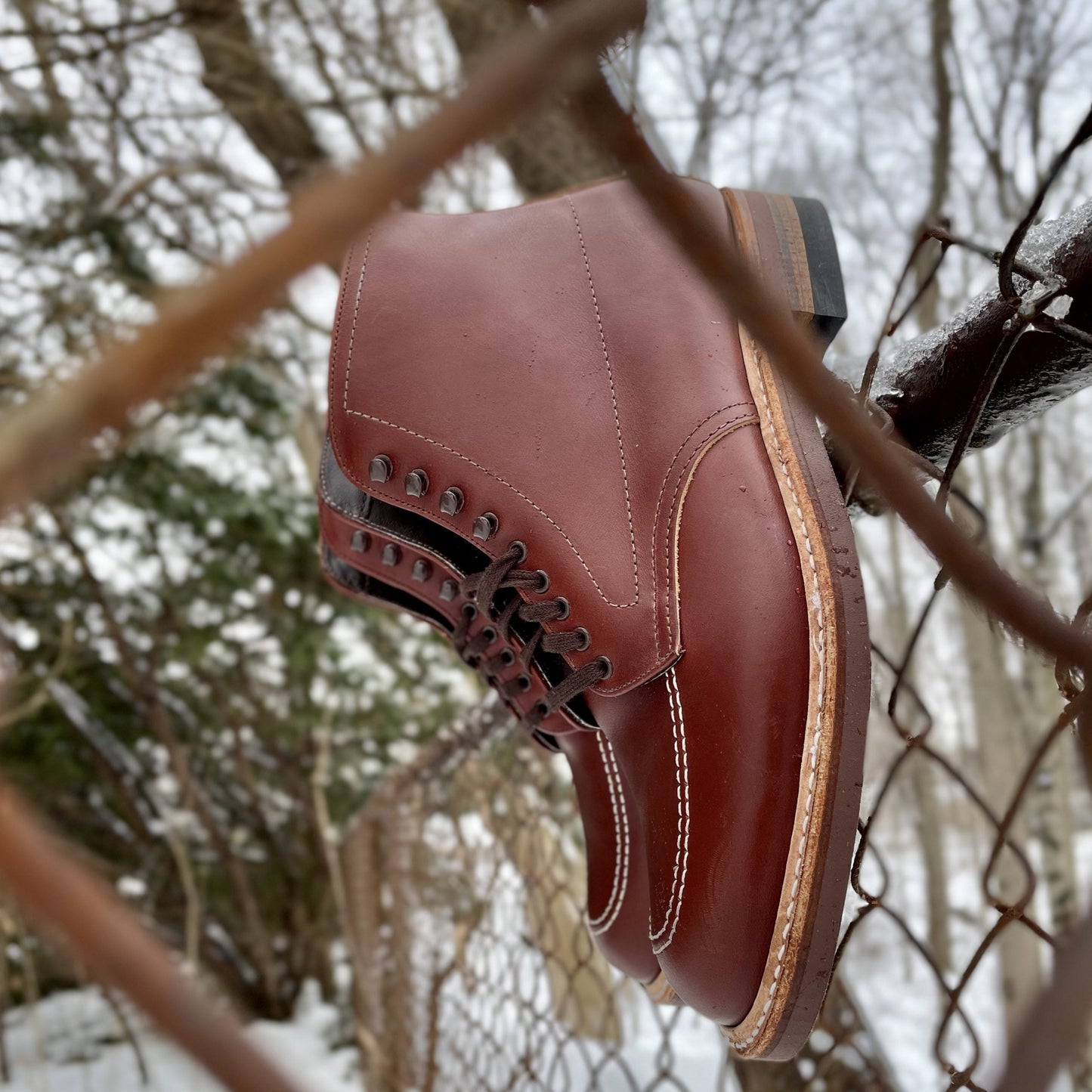 A close-up of the Alden Madison 405 - Indy Boot in mid-brown calfskin, featuring detailed stitching and sleek black eyelets, rests on a rusted chain-link fence. It creates a rustic winter scene against bare trees and snowy landscape.