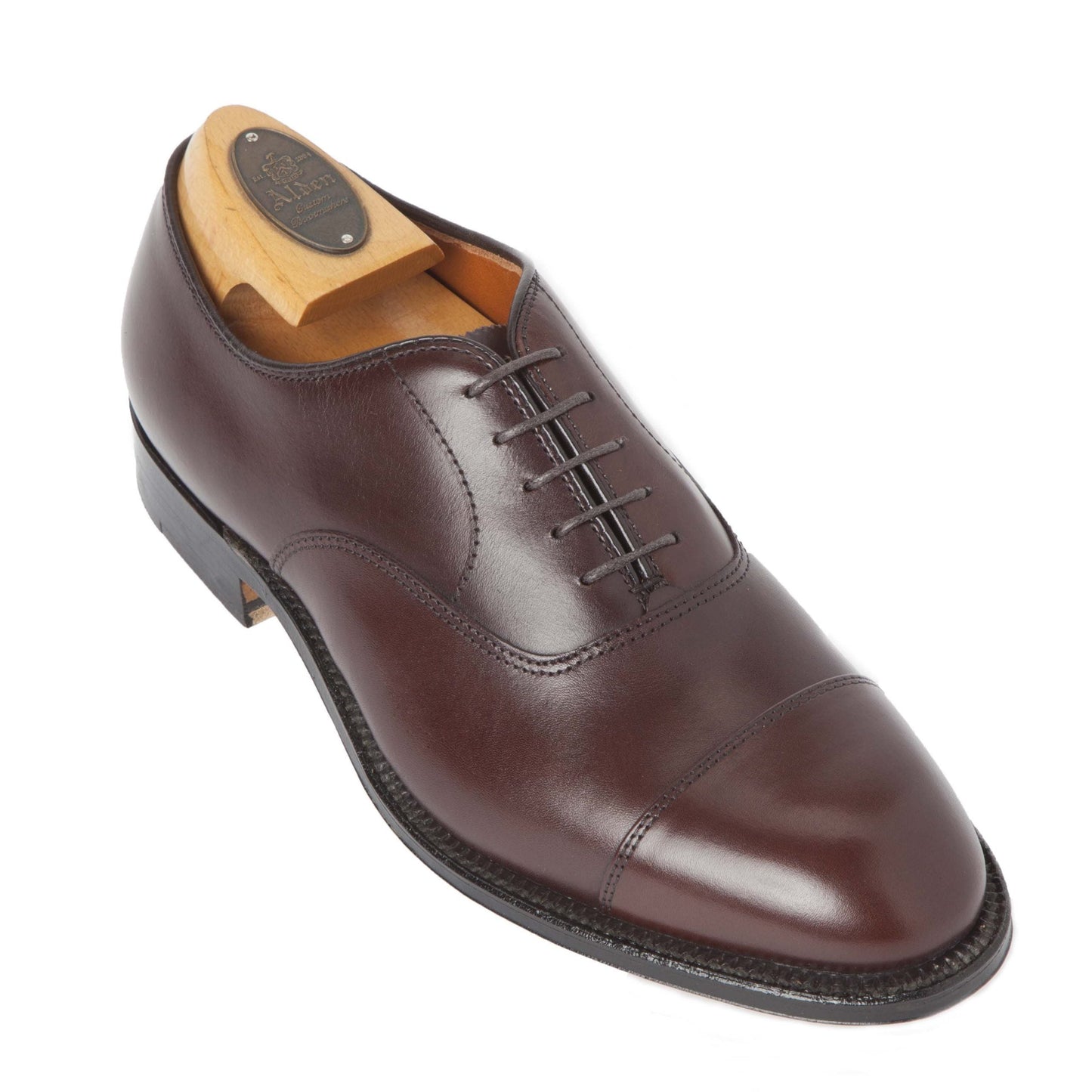 The Alden Madison 920 Cap Toe Bal Oxford, crafted in polished brown calfskin, showcases the Hampton Last for a refined fit. It features an elegant cap toe design and leather sole, with a wooden shoe tree inside. A small engraved oval plate adorns it, viewed at a slight angle.