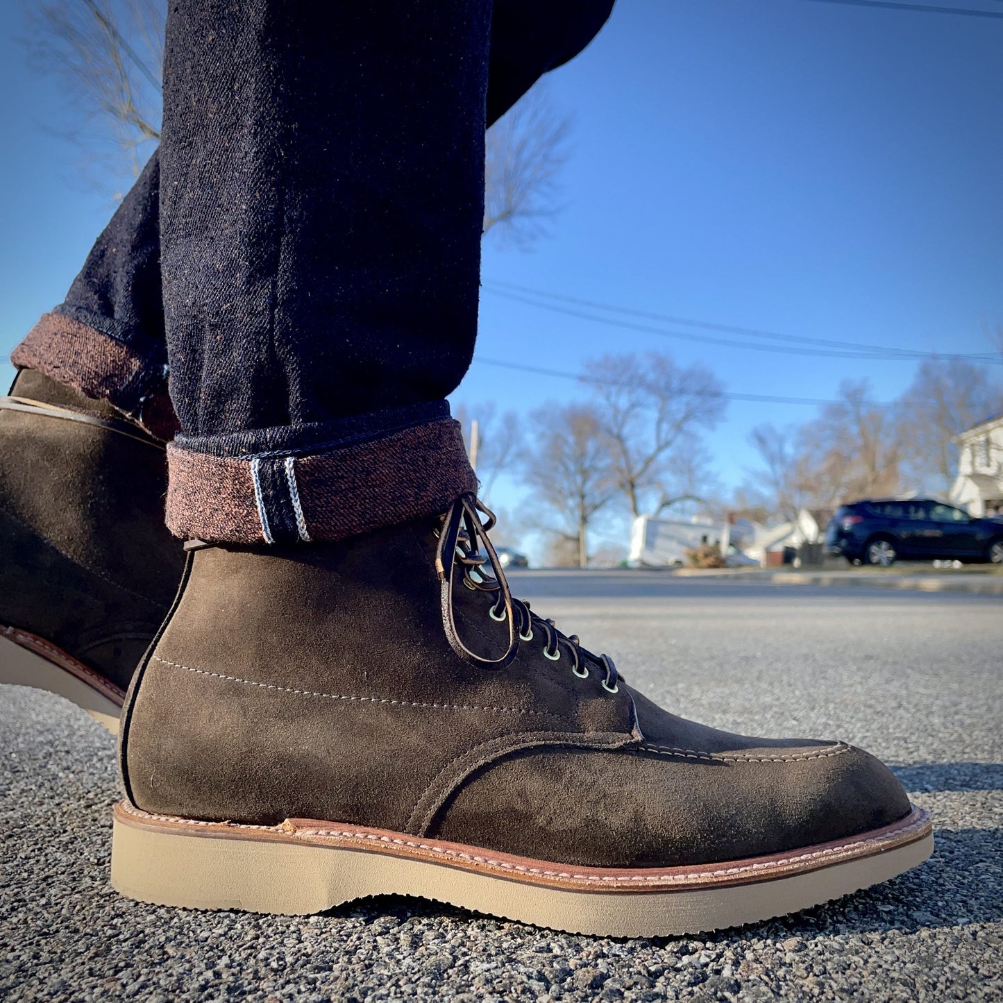 A close-up shows a person wearing Alden Madisons D0938H - Duncan Indy Boot in Loden Suede. The boots feature laces, contrast stitching, and light brown soles, paired with dark cuffed jeans on a paved surface under a clear blue sky with trees and a parked car in the background.