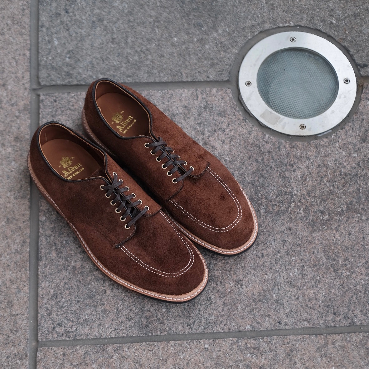 The Alden Madison D2604C Indy Shoe in Tobacco Reverse Chamois, crafted on the Trubalance Last with white stitching and dark laces, elegantly placed on stone pavement, is accentuated by a circular metallic light fixture.