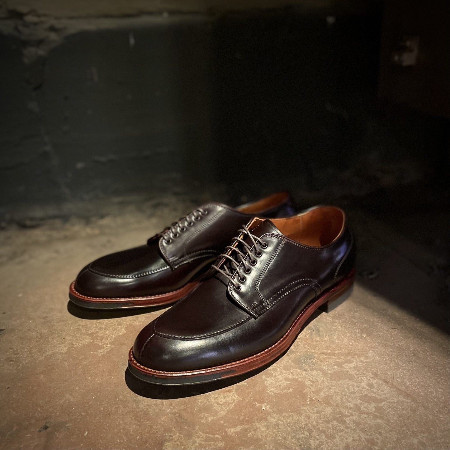 A dimly lit scene features Alden Madisons D1629C - V Tip Split Toe Blucher in Color 8 Shell Cordovan leather with red-brown soles on a concrete floor. Shadows accentuate their elegance against a textured dark wall in the background.
