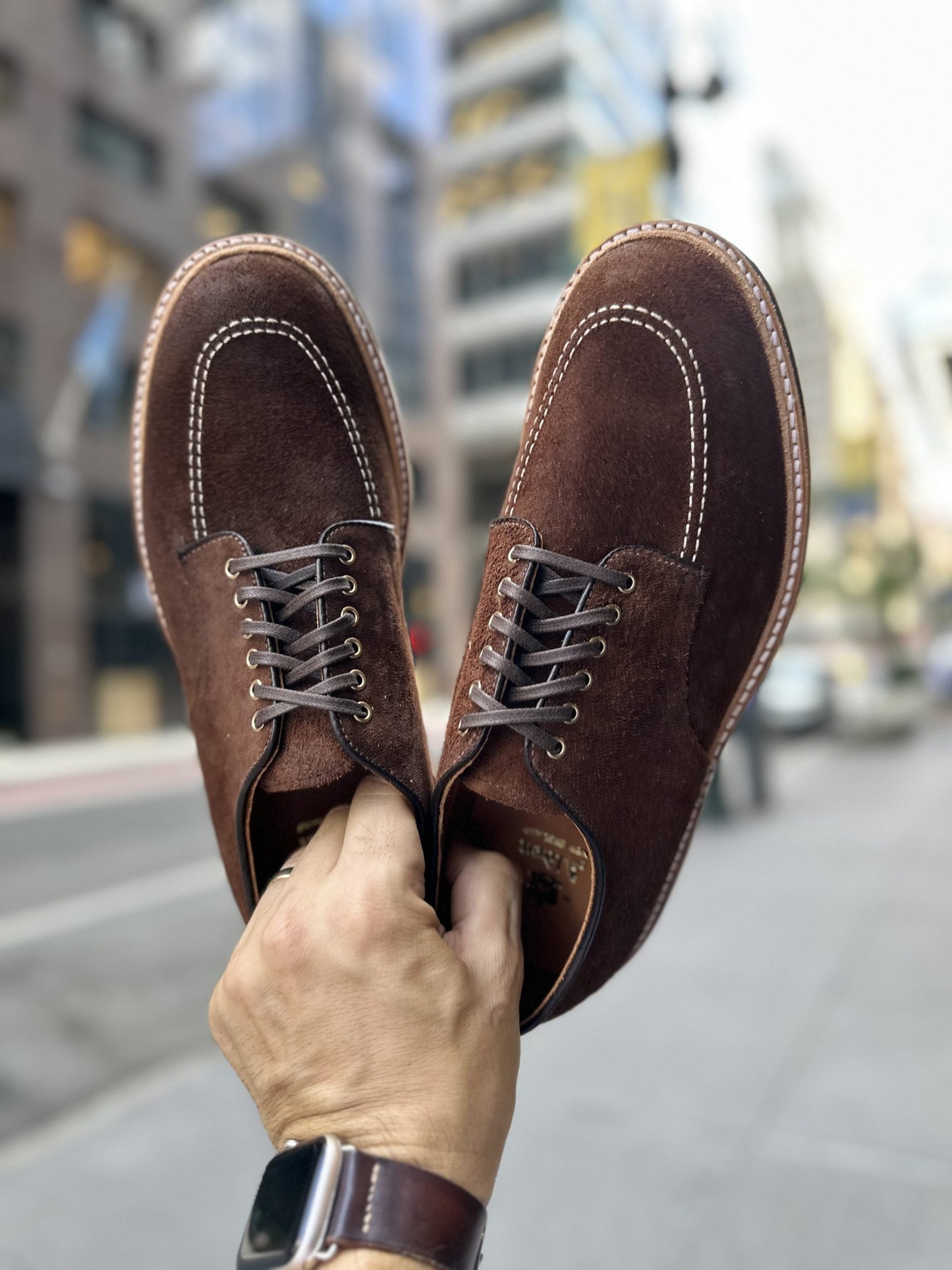 In an urban setting, someone holds the Alden Madison D2604C - Indy Shoe in Tobacco Reverse Chamois, featuring standout white stitching. The blurred background reveals city buildings and streets, while a watch with a brown leather strap adorns the persons wrist.