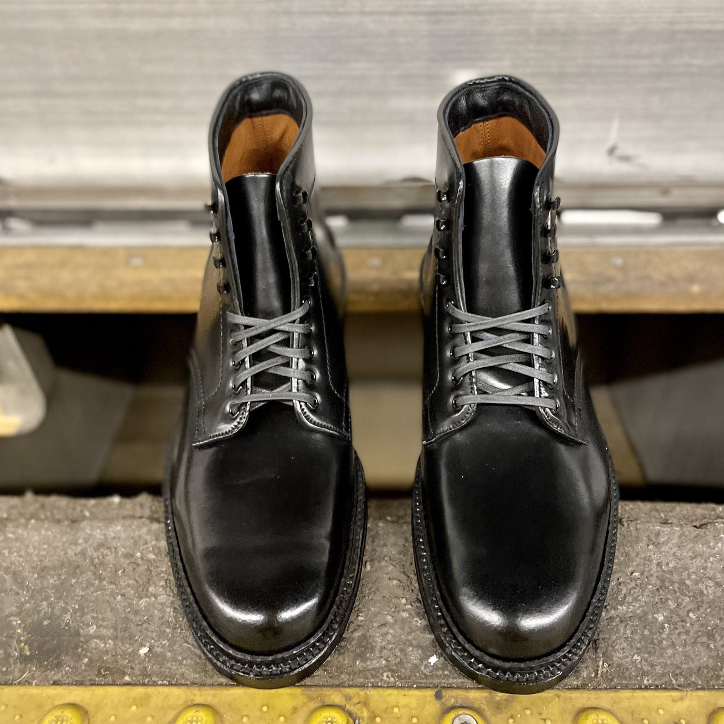 The Alden Madison Director boots, model D6842H in Black Shell Cordovan, feature sleek shiny leather and gray laces with speed hooks. These stylish boots, with a double oak leather sole, are placed together on a textured surface near a metallic background.