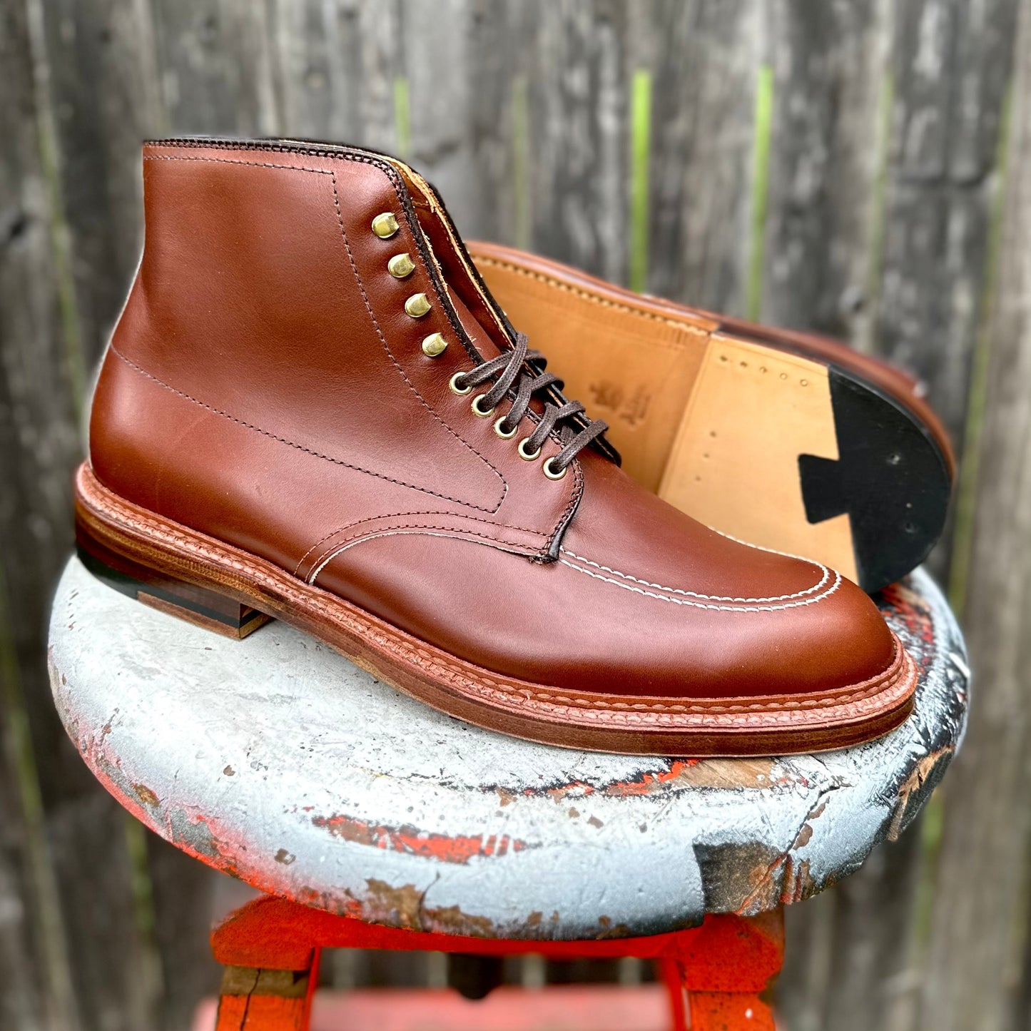 The D2940H - Madison Indy Boot in Madison Brown Calf by Alden Madison, featuring polished leather, detailed stitching, and brass eyelets, rests on a weathered stool. Its contrasting dark sole and heel enhance the style against a rustic wooden fence backdrop.