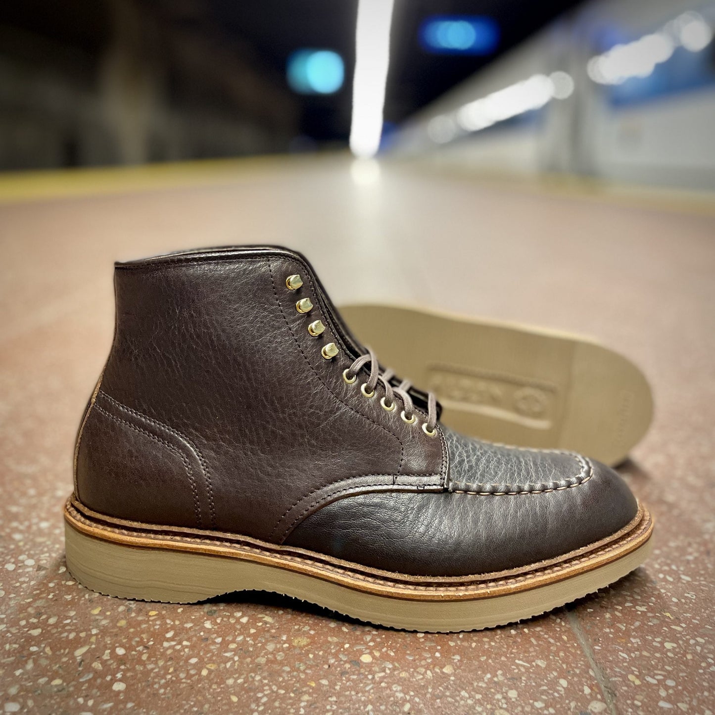 An Alden Dirty Bones Indy Boot 2.0, crafted in Arabica Lux leather, rests on a subway platform with its sole visible. The blurred background of a train and platform lights emphasizes the contrast between the rugged boot and its urban surroundings.