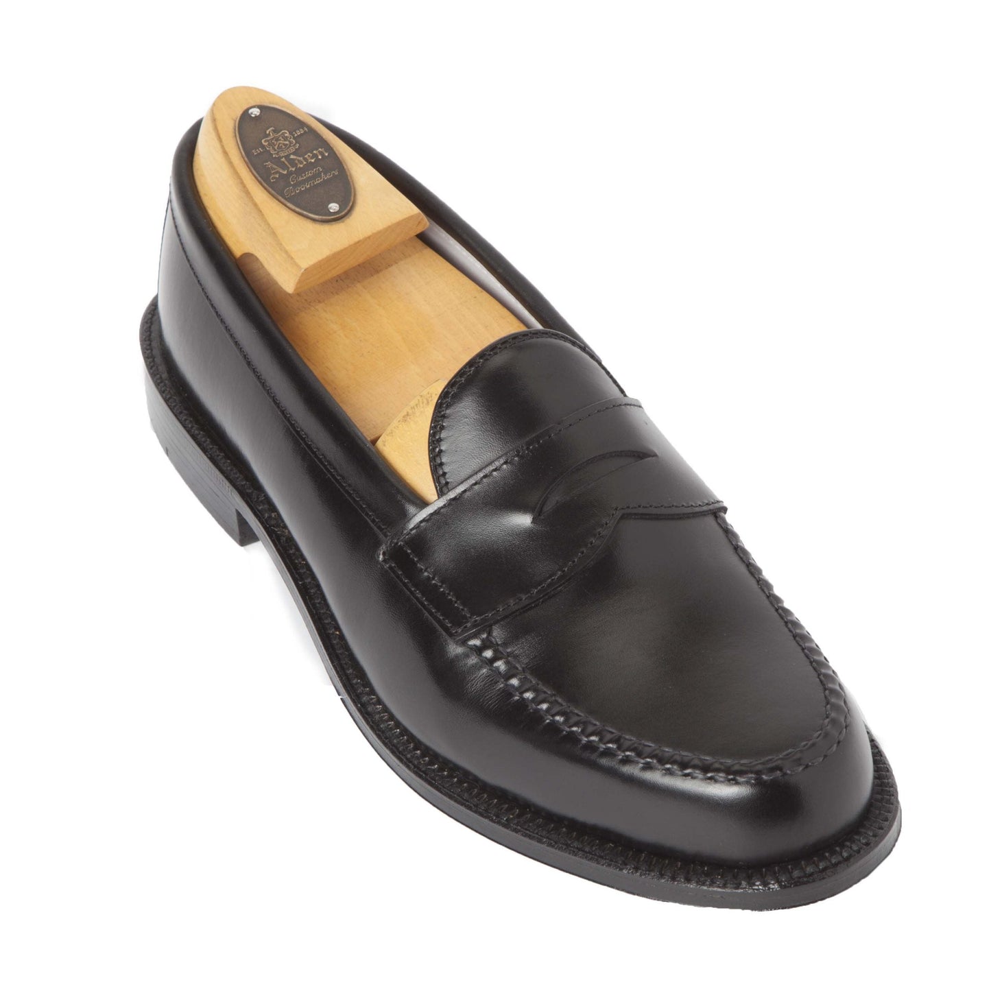 A black calfskin loafer, 981 - Leisure Hand Sewn by Alden Madison, is showcased on a white background. It boasts a classic penny loafer design with a wooden shoe tree inside, featuring an engraved oval plaque reading Alden. The leather outsole enhances its elegance.