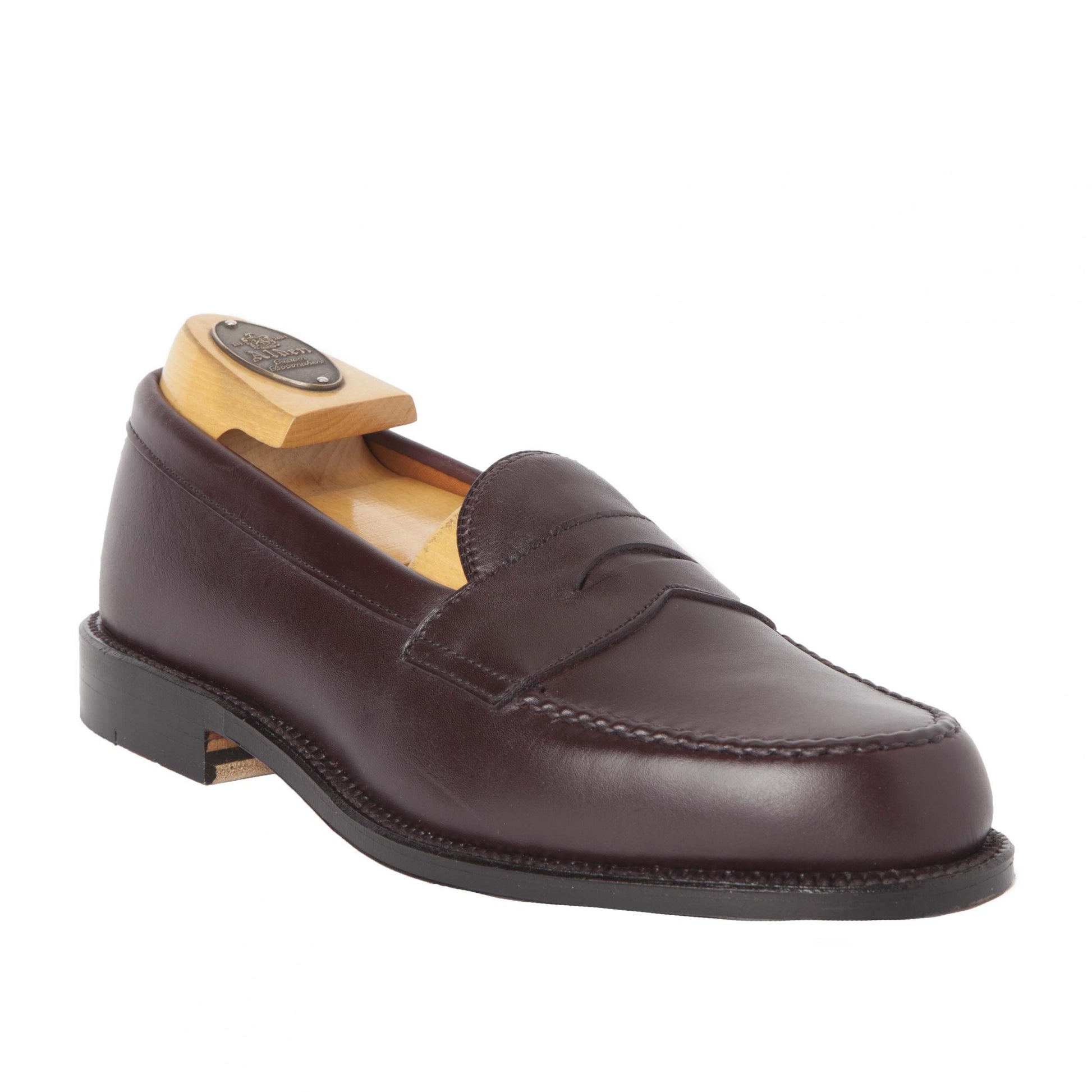 A single burgundy calfskin loafer by Alden Madison, crafted in a Van Last silhouette, is displayed at an angle with a wooden shoe tree. Featuring a penny slot, it has an oak leather sole and polished finish for timeless elegance. Product Name: 984 - Leisure Hand Sewn.