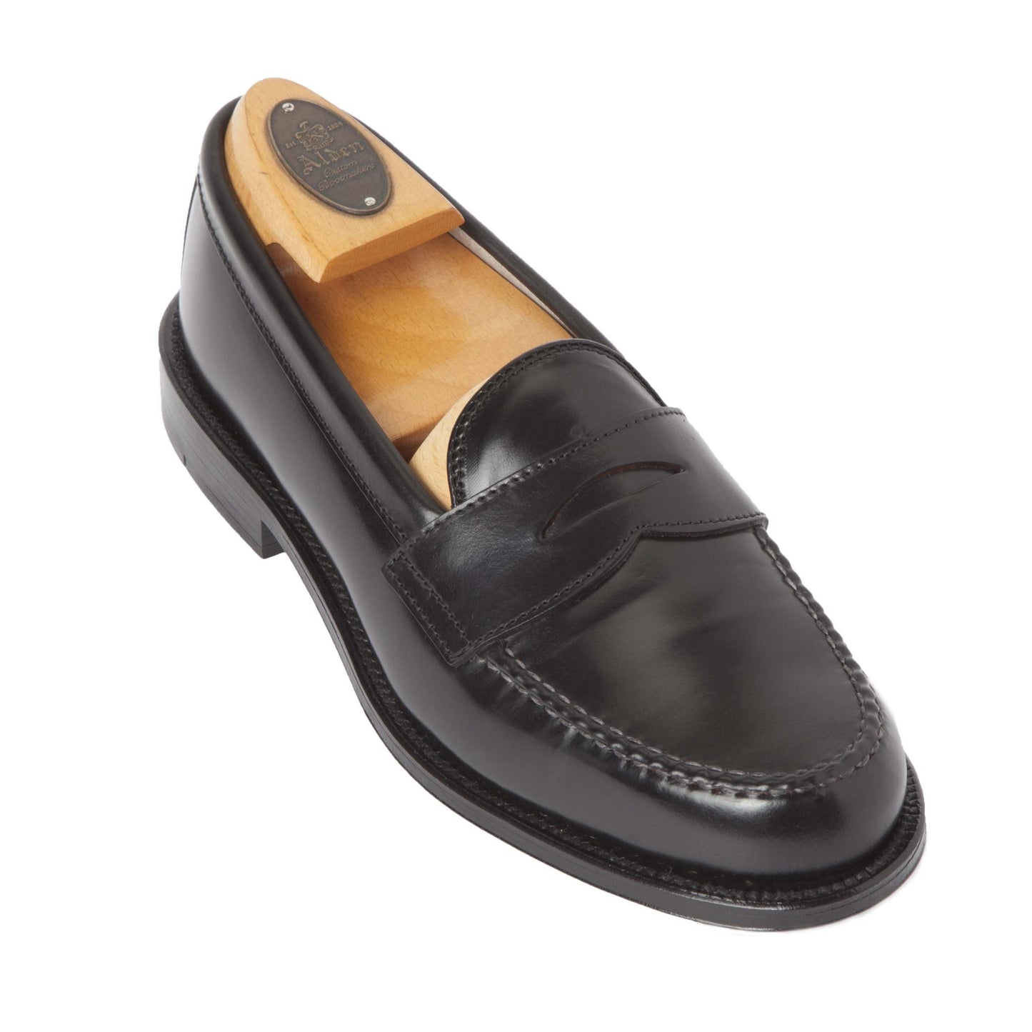 A black Shell Cordovan loafer by Alden Madison (Product 987 - Leisure Hand Sewn) is shown with a wooden shoe tree on a white background. It features a classic penny slot and visible apron stitching, with a polished finish.