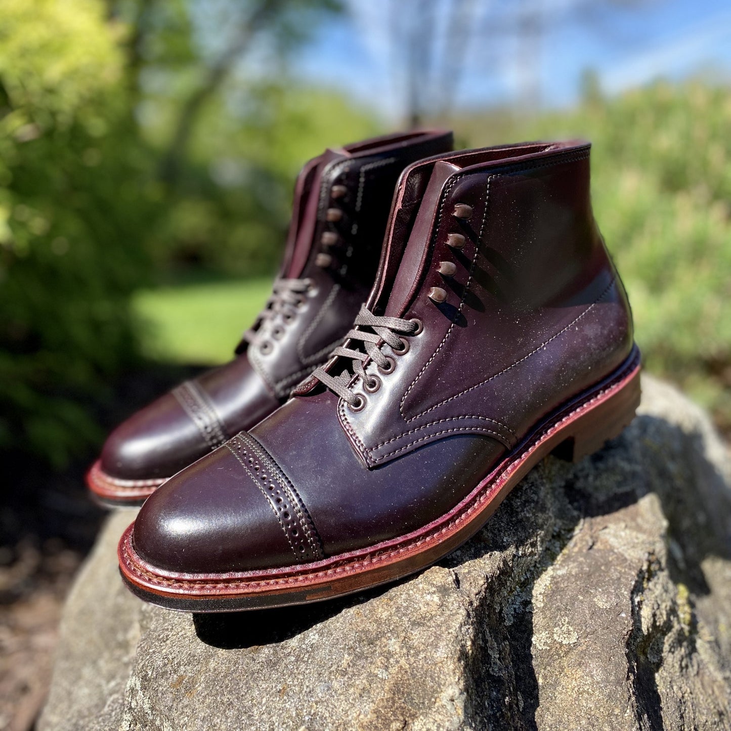 A pair of Alden Madisons D6861HC - Jumper Boot in Color 8 Shell Cordovan, featuring polished brown leather and a contrasting light brown sole, sits on a rock. These cap-toe Barrie Last boots are outdoors against blurred greenery.