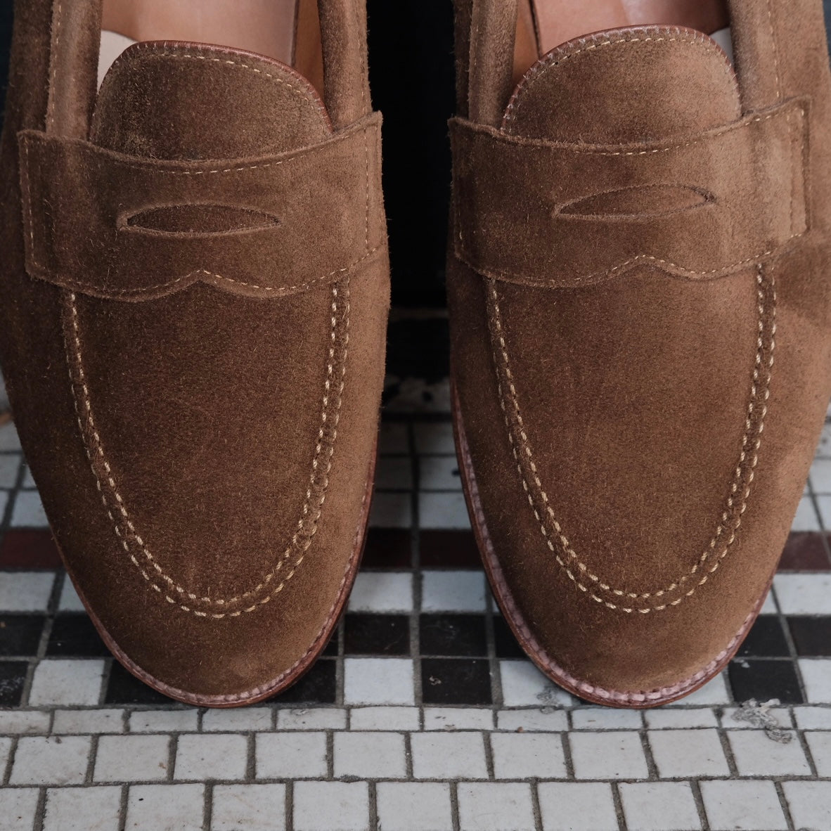 Close-up of Alden Madisons 96941F - Flex Penny Loafer in Snuff Suede on a patterned black, white, and gray tiled floor, highlighting the detailed stitching and craftsmanship.