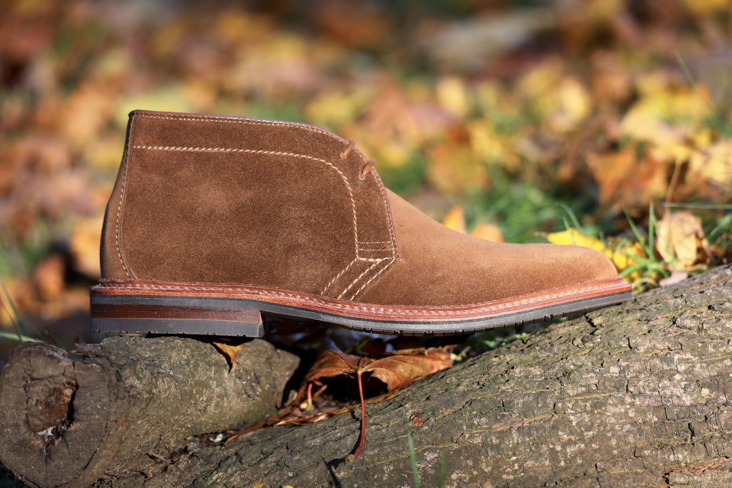 A snuff suede chukka boot by Alden Madison, model 1593L with an L3 sole, rests on a tree trunk amidst autumn leaves. Its intricate stitching and rich texture are illuminated in the sunlight.