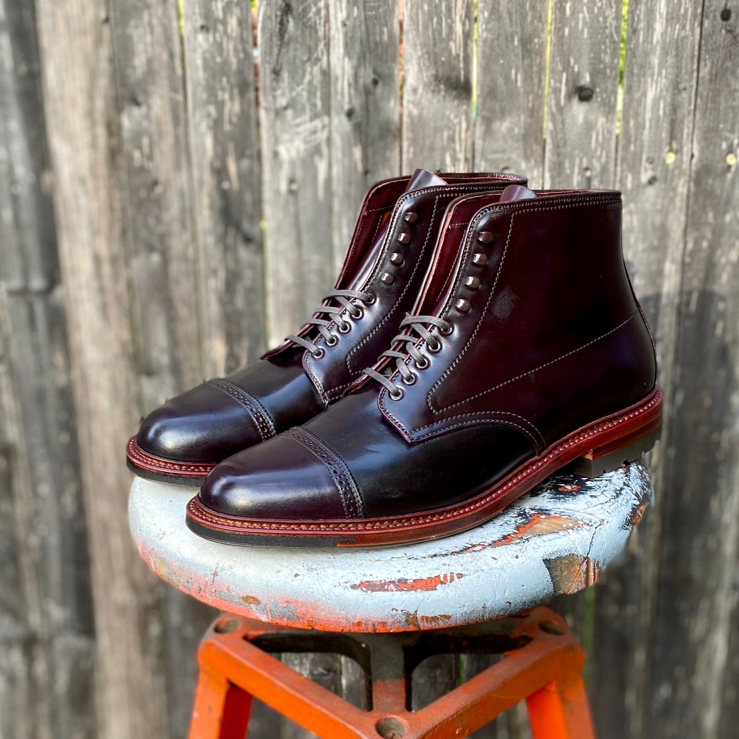 The D6861HC - Jumper Boot in Color 8 Shell Cordovan by Alden Madison, crafted on the Barrie Last with cap toes, contrasting stitching, and a Commando Sole, rests on a rustic stool against a weathered wooden backdrop.
