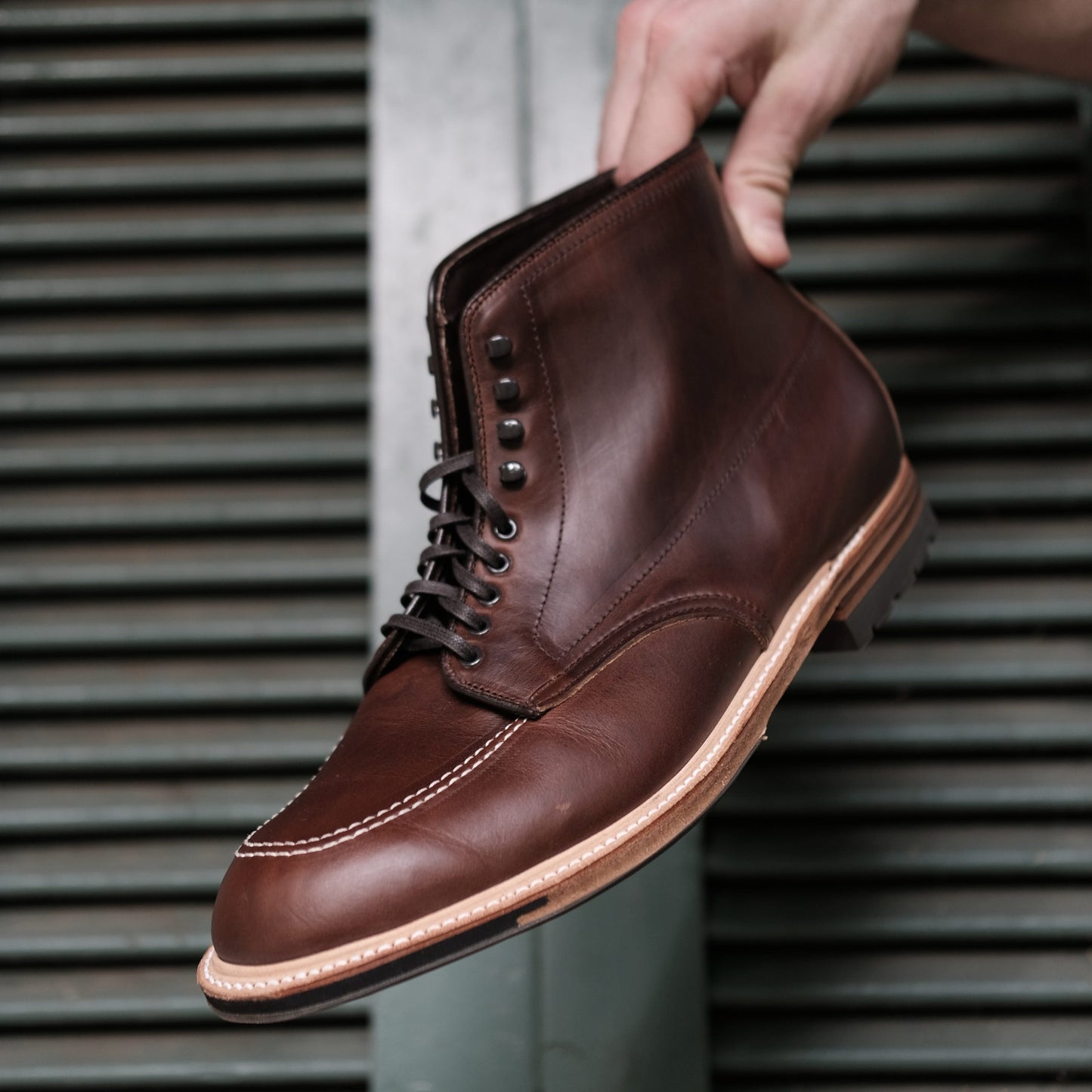 A hand holds an Alden Madison 403C Indy Boot with a Commando Sole against metal slats, featuring Brown Chromexcel leather on the Truebalance Last, a round toe, contrast stitching, and multiple metal eyelets for lacing.