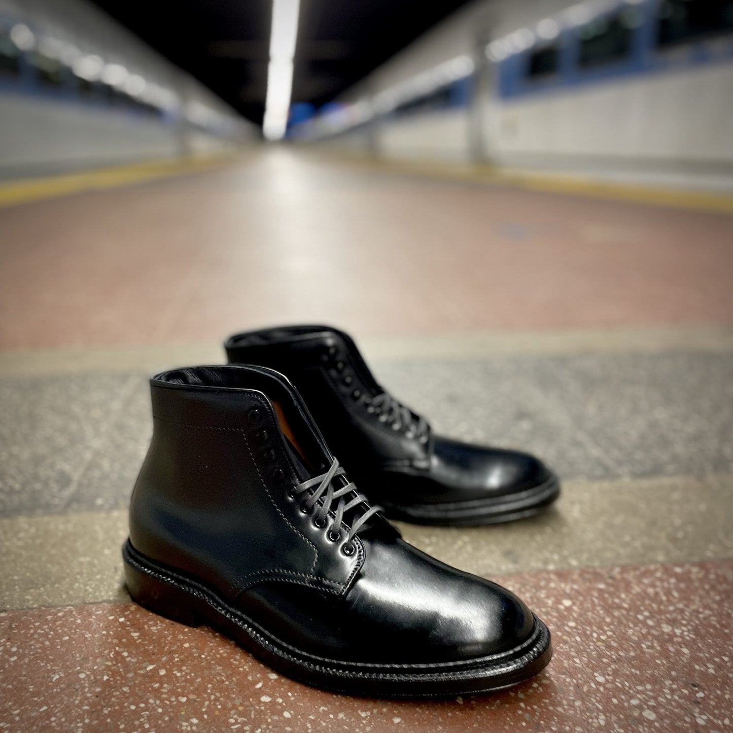 In an empty subway station, the D6842H - Plain Toe Director Boot in Black Shell Cordovan from Alden Madison sits on a speckled floor. The polished boots with speed hooks and laces stand against a blurred background, highlighting their elegance and featuring a Double Oak Leather Sole.