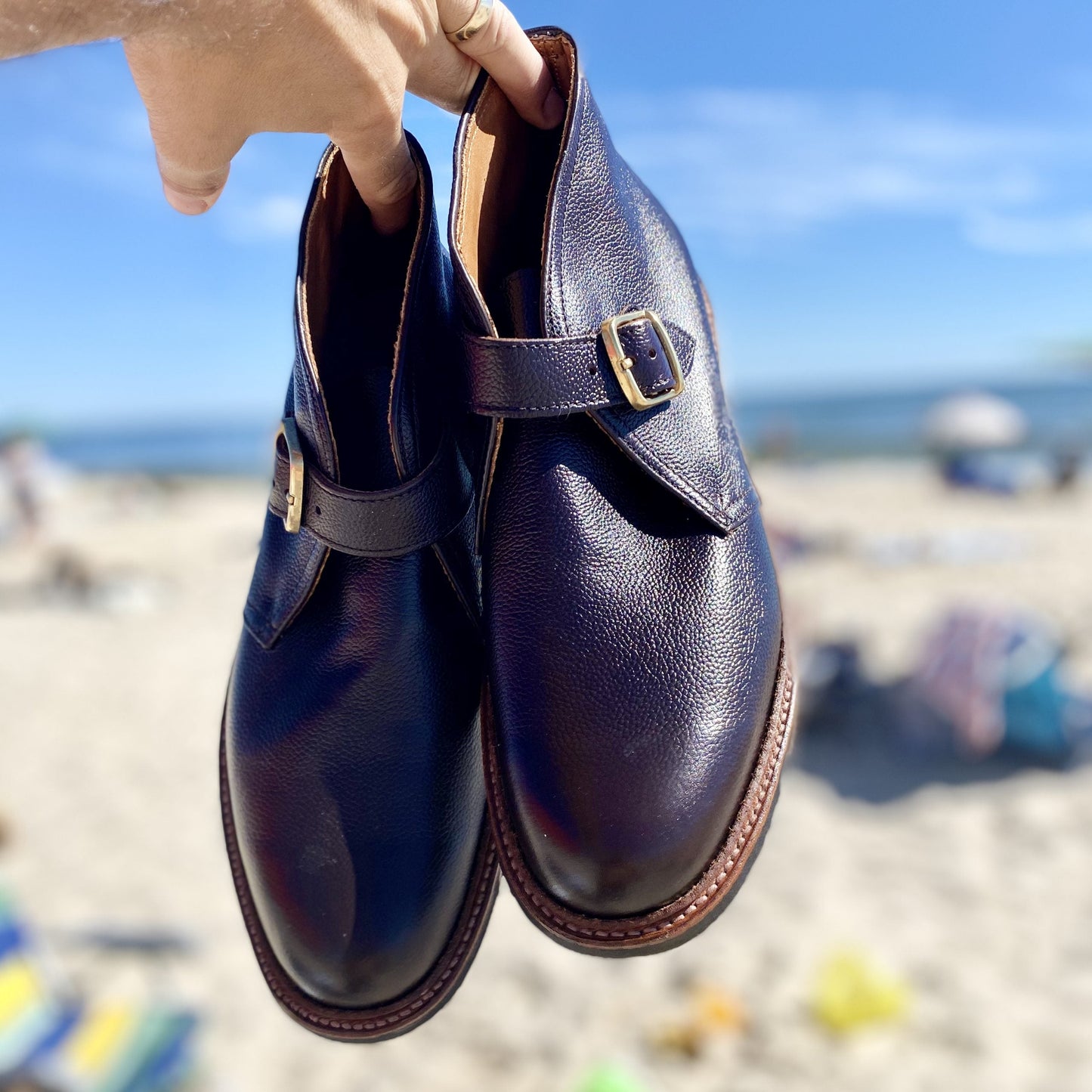 The Alden Madison D9704C - George Boot in Brown Regina Grain is showcased against a blurred beach, with sand meeting the ocean under a clear blue sky.