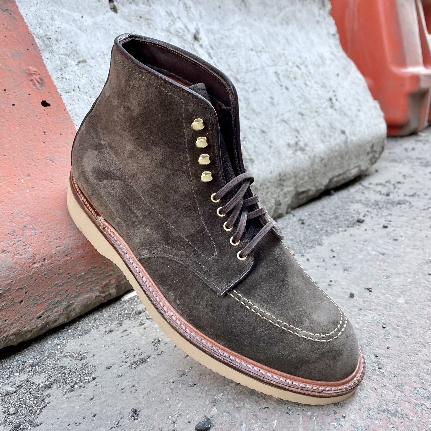 A D0938H - Duncan Indy Boot in Loden Suede by Alden Madison, featuring gold eyelets and brown laces, rests against a light gray and orange concrete barrier. The beige sole complements the rugged aesthetic on a rough surface.