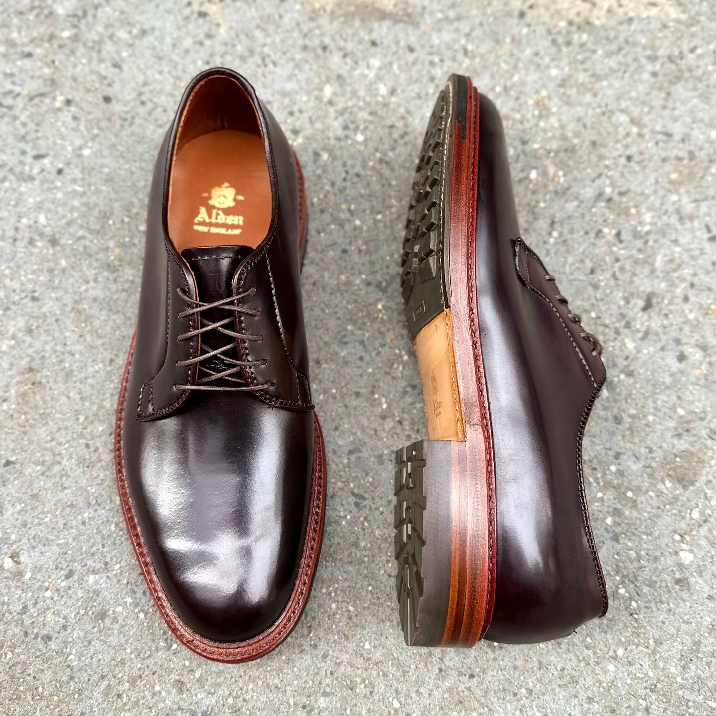 A pair of Alden Madison D7406C Antique Plain Toe Blucher shoes in Color 8 Shell, featuring red soles, is displayed on a concrete surface. One shoe is upright showing the top, and the other lies on its side to highlight the Barrie Last construction and heel detail.