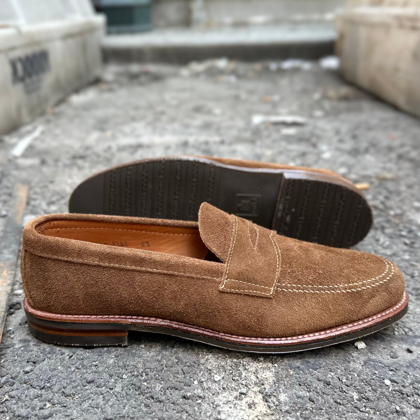 Alden Madisons 6221L - Unlined Penny Mocc in Snuff Suede features a moc toe and visible stitching with a penny slot on the vamp. Grappling with gravel, one shoe highlights its rubber sole, while the other reveals textured detail.