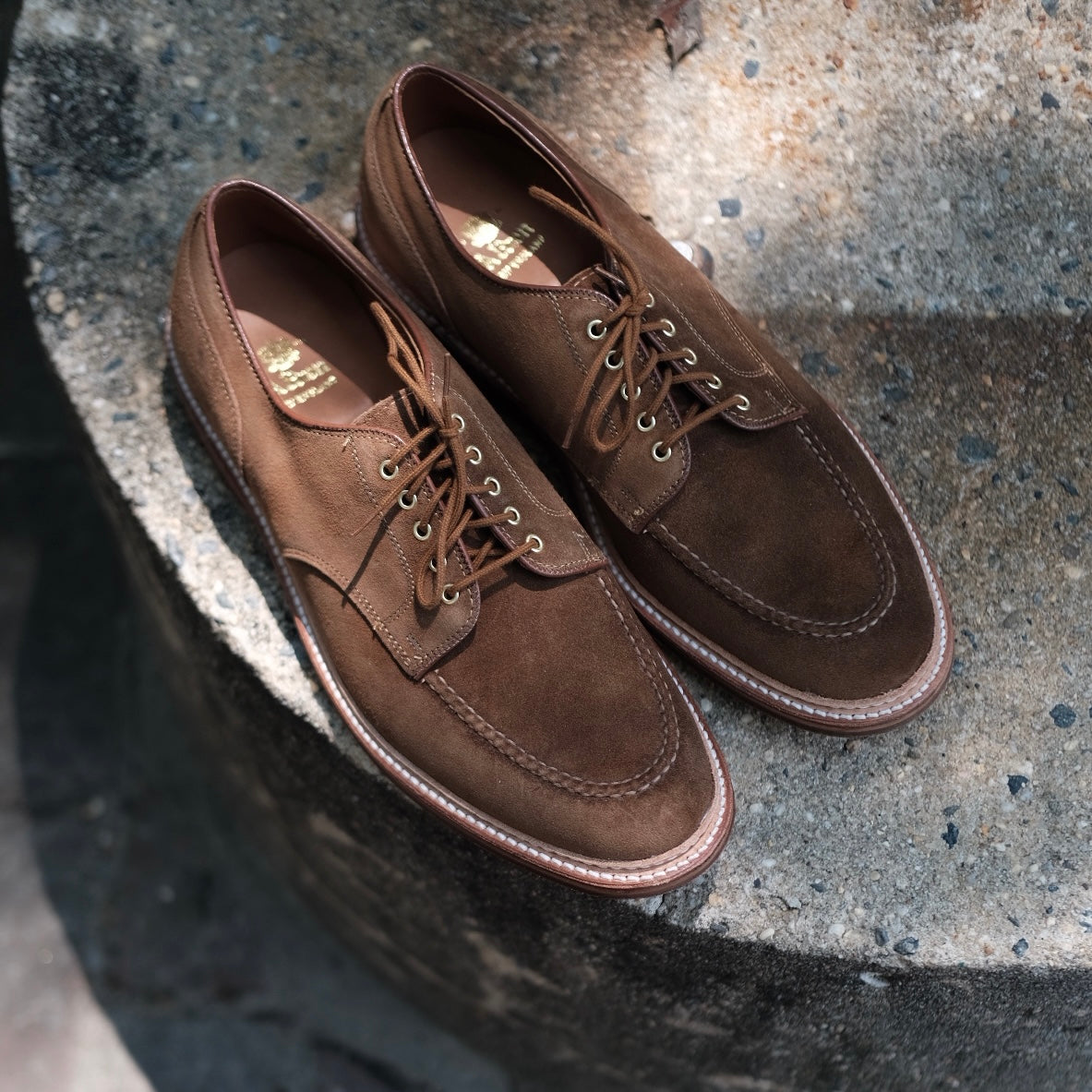 The Quickley in Snuff Suede by Alden Madison features brown suede lace-up shoes on textured concrete; crafted in the classic Barrie Last style, their snuff suede finish glows in natural light, casting soft elegant shadows.