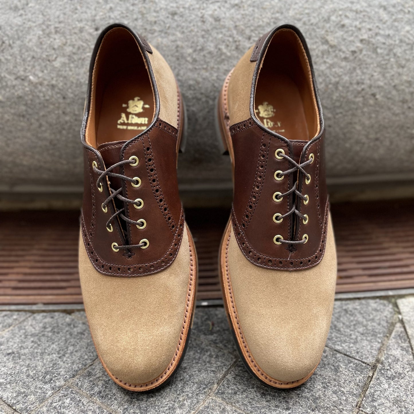 The Alden Madison D1304C - Valhalla Saddle Oxford in CXL and Tan Suede showcases dark brown leather around the laces, tan suede on the toe and sides, a durable commando sole, and laces up elegantly on a stone surface.
