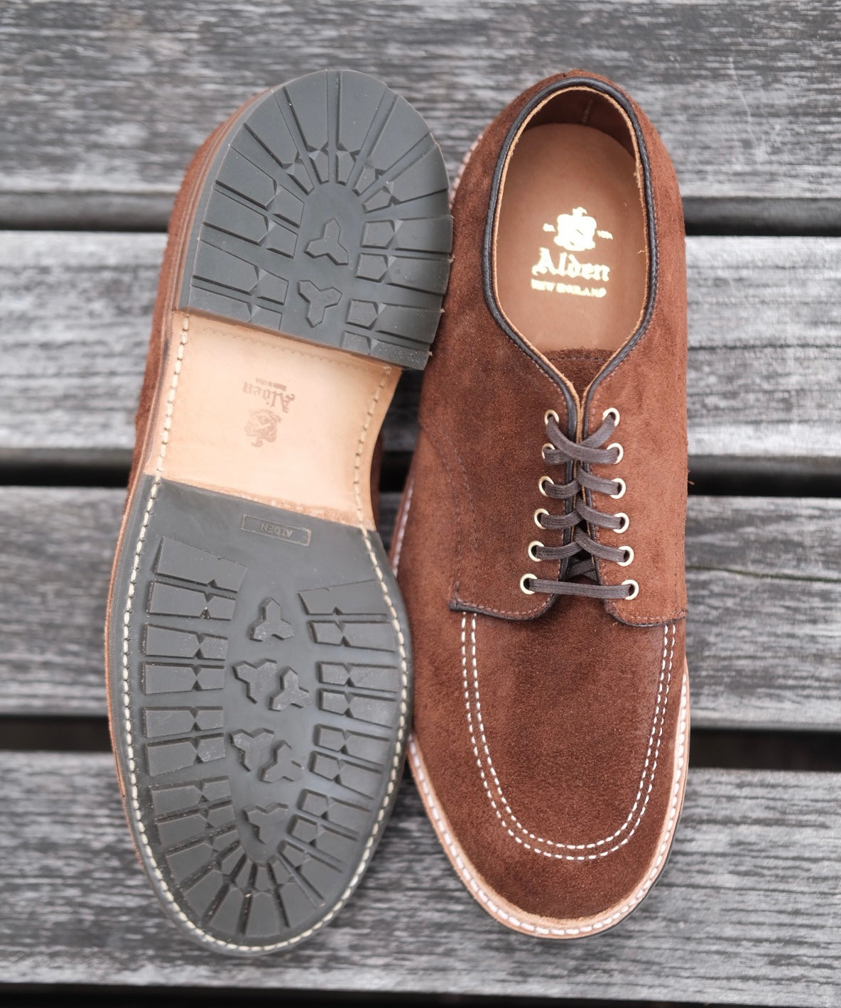 The Tobacco Reverse Chamois Indy Shoes by Alden Madison are displayed on a wooden surface. One shoe stands upright, highlighting its laced design and Trubalance Last, while the other lies on its side to show the tread pattern. Alden New England is printed on the insole.