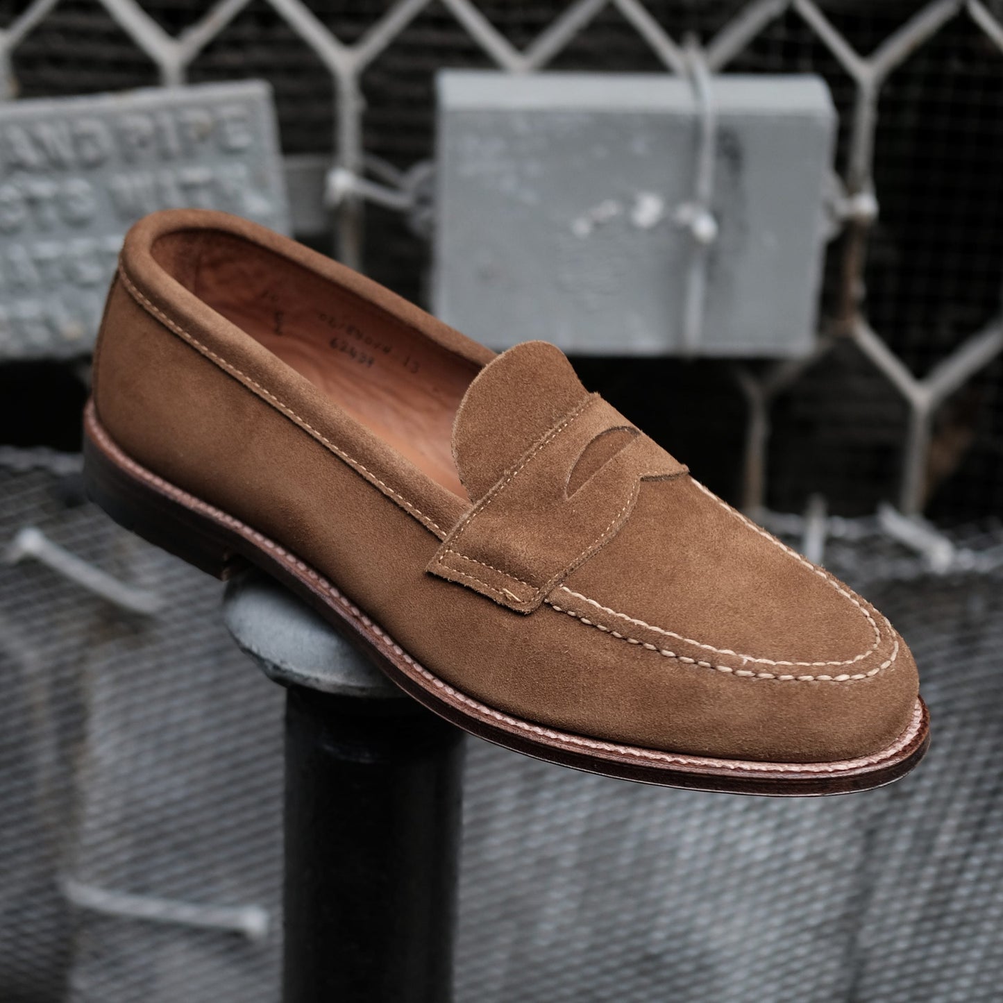 A single Alden Madison 6243F loafer in snuff suede, featuring a penny slot and white stitched sole, is poised on a black metal post against a chain-link fence and blurred signs.