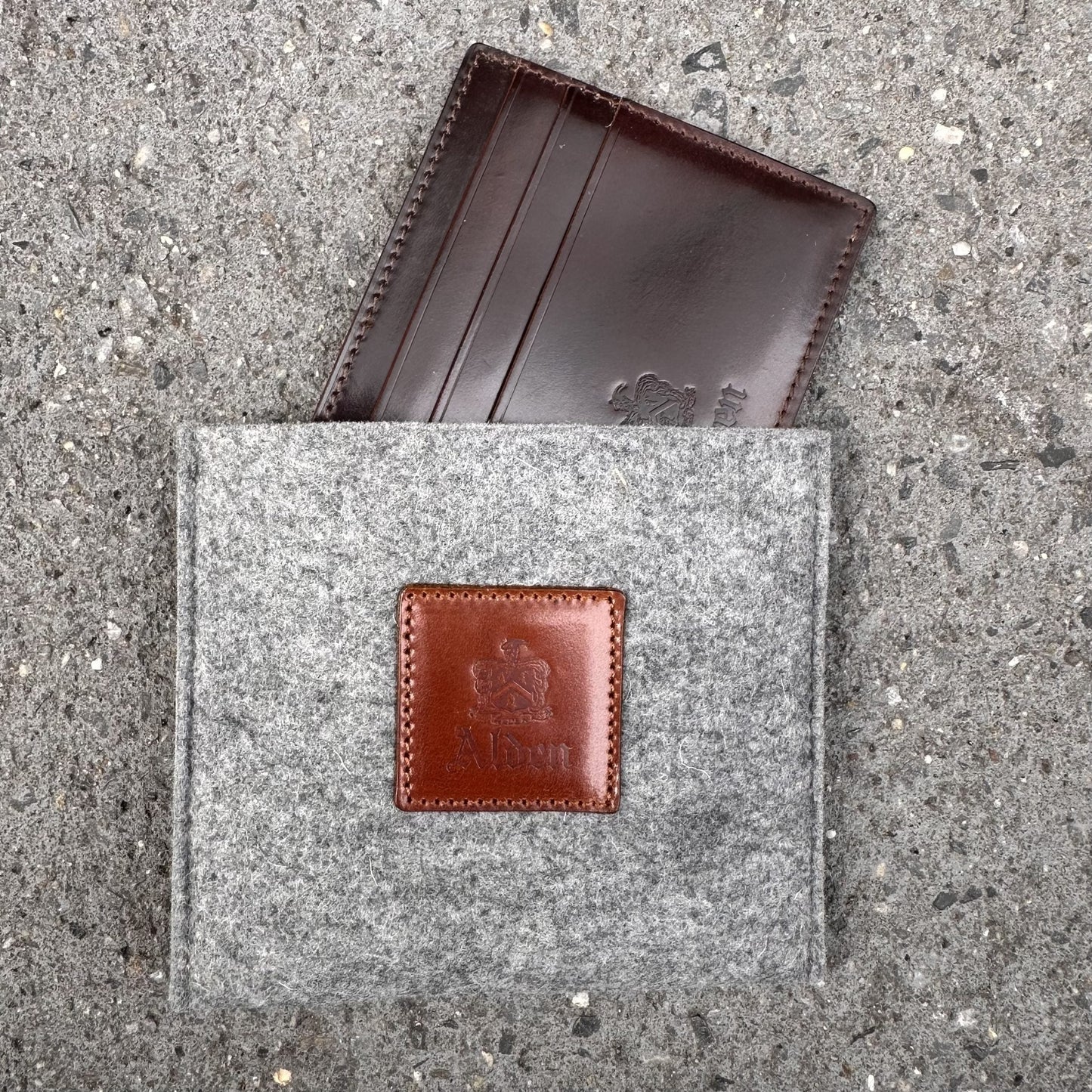The LG 6008 - Alden Color 8 Shell Credit Card Holder by Alden Madison, a brown leather cardholder with 6 pockets, slides into a gray felt pouch featuring a front leather patch. Both items showcase embossed logos against a speckled concrete backdrop, enhancing the urban style of this accessory set.