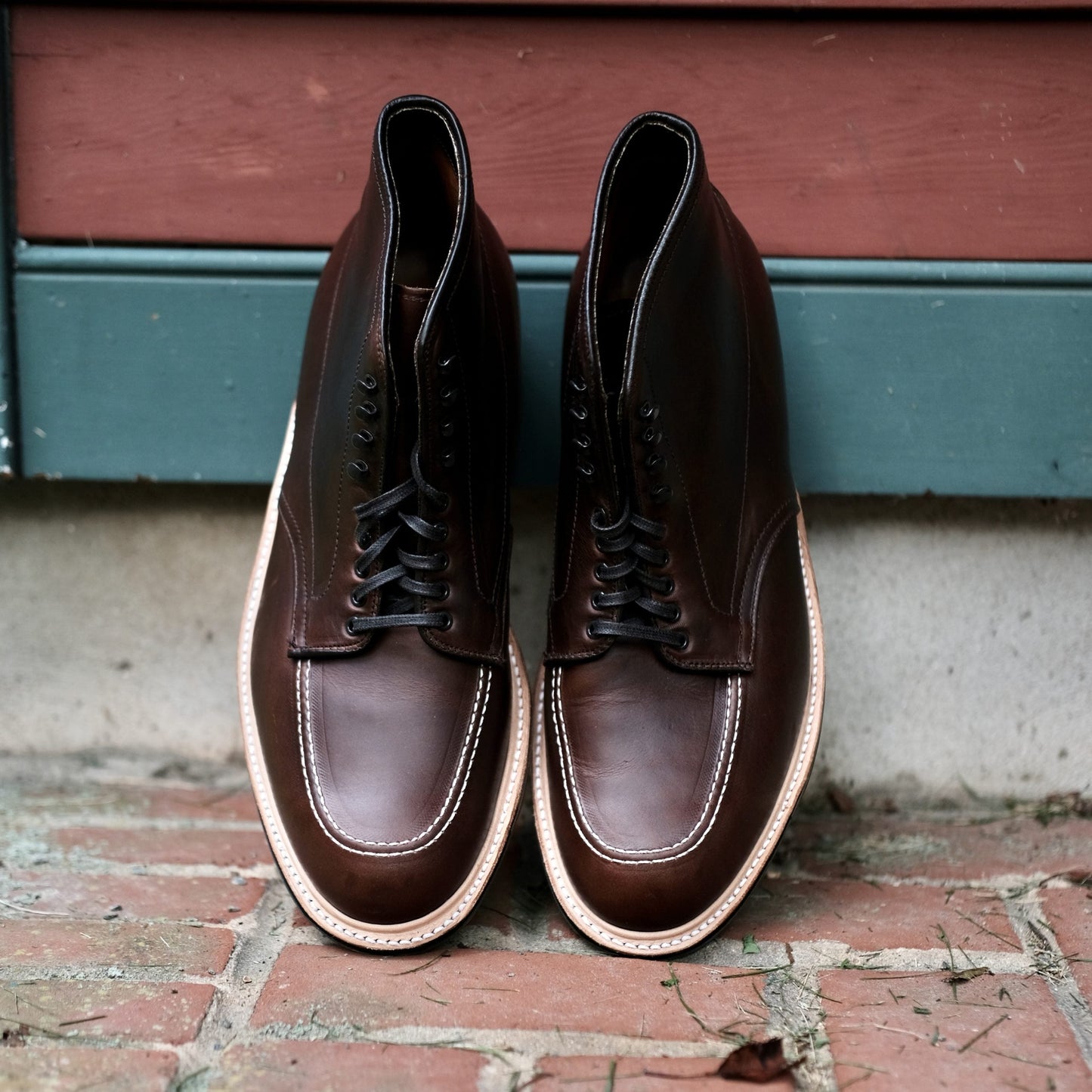 A pair of Alden Madison 403C Indy Boots in dark brown Chromexcel leather, featuring light Commando soles and visible toe stitching on the Truebalance Last design for comfort, rests side by side on a brick surface against a painted wooden wall.