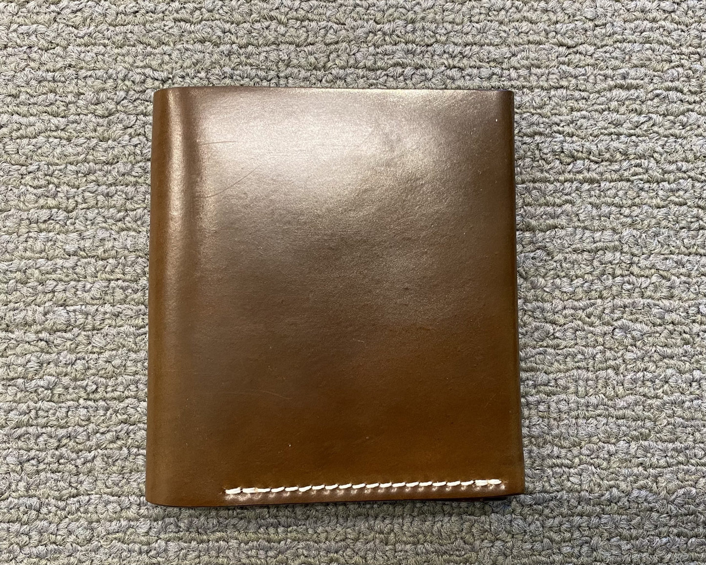 The LG 7007 - Alden Cognac Shell Cordovan Wallet by Alden Madison, with a closed brown leather design and white stitching, sits elegantly on a gray textured surface.