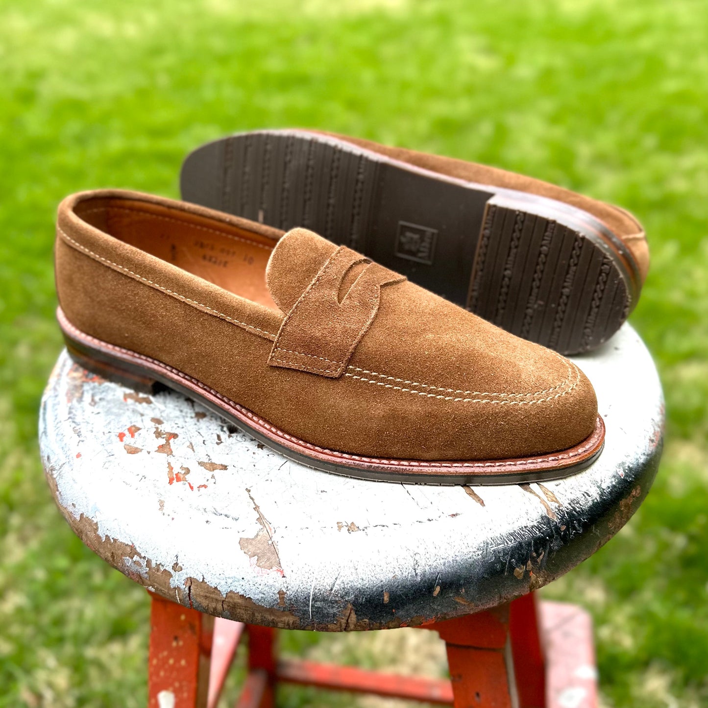 A pair of Alden Madisons 6221L - Unlined Penny Mocc in Snuff Suede, featuring a Mocc Toe design and visible stitching, sits on a weathered stool with a blurred grass backdrop. One shoe stands upright while the other tilts to display its durable rubber sole, exuding effortless style.