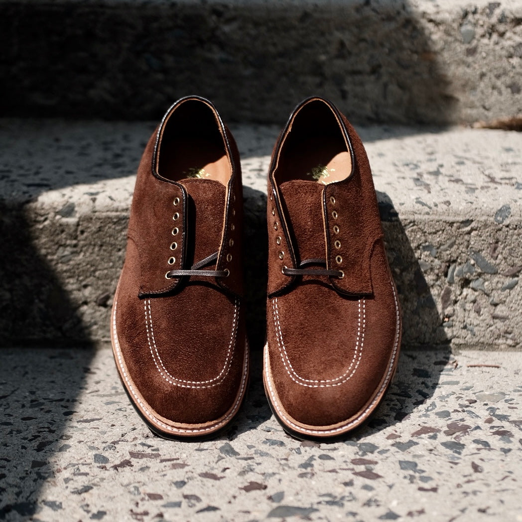 The D2604C Indy Shoe in Tobacco Reverse Chamois by Alden Madison features brown suede, white stitching, black laces, and a rounded toe designed on the Trubalance Last. They rest stylishly on coarse concrete steps under sunlight.