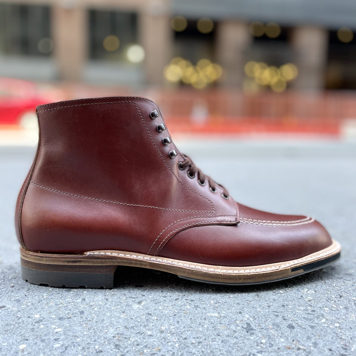 Close-up of the Alden Madison 405C - Original 405 Indy Boot in brown leather with black laces and a commando sole, resting on gray concrete. Blurred city buildings and lights create a softly illuminated background.