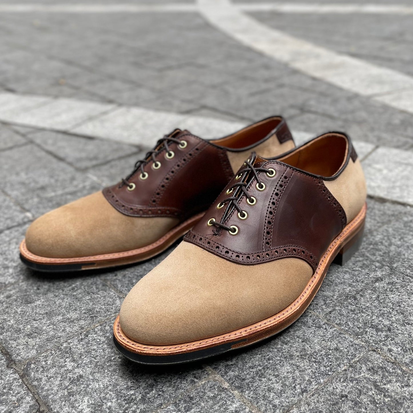 The Valhalla Saddle Oxford in CXL and Tan Suede by Alden Madison, featuring black laces and a natural Commando sole, rests elegantly on gray stone pavement with a light pattern.