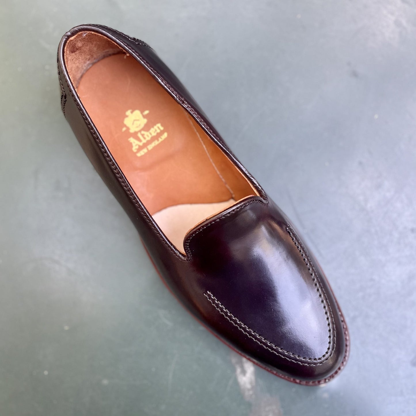 A top view of the D1118 - Vanderbilt Slip On in Color 8 Shell by Alden Madison reveals a dark brown leather loafer made of Shell Cordovan. It has a tan interior with Alden and New England, built on an Aberdeen Last, sitting on a subtly textured gray surface, highlighting its Leather Sole.