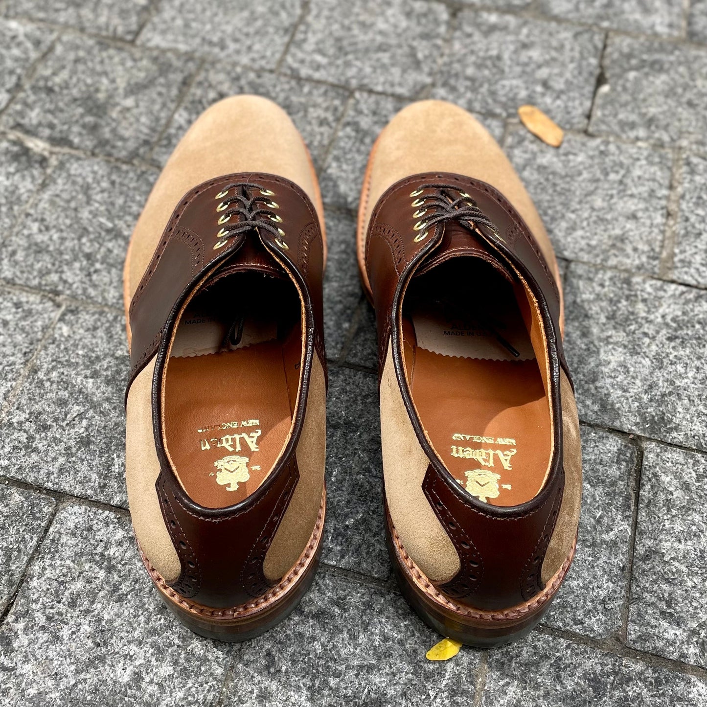 A pair of D1304C - Valhalla Saddle Oxford shoes in CXL and tan suede by Alden Madison, featuring visible brand markings inside and brown laces, rests on a stone pavement with a small yellow leaf nearby.