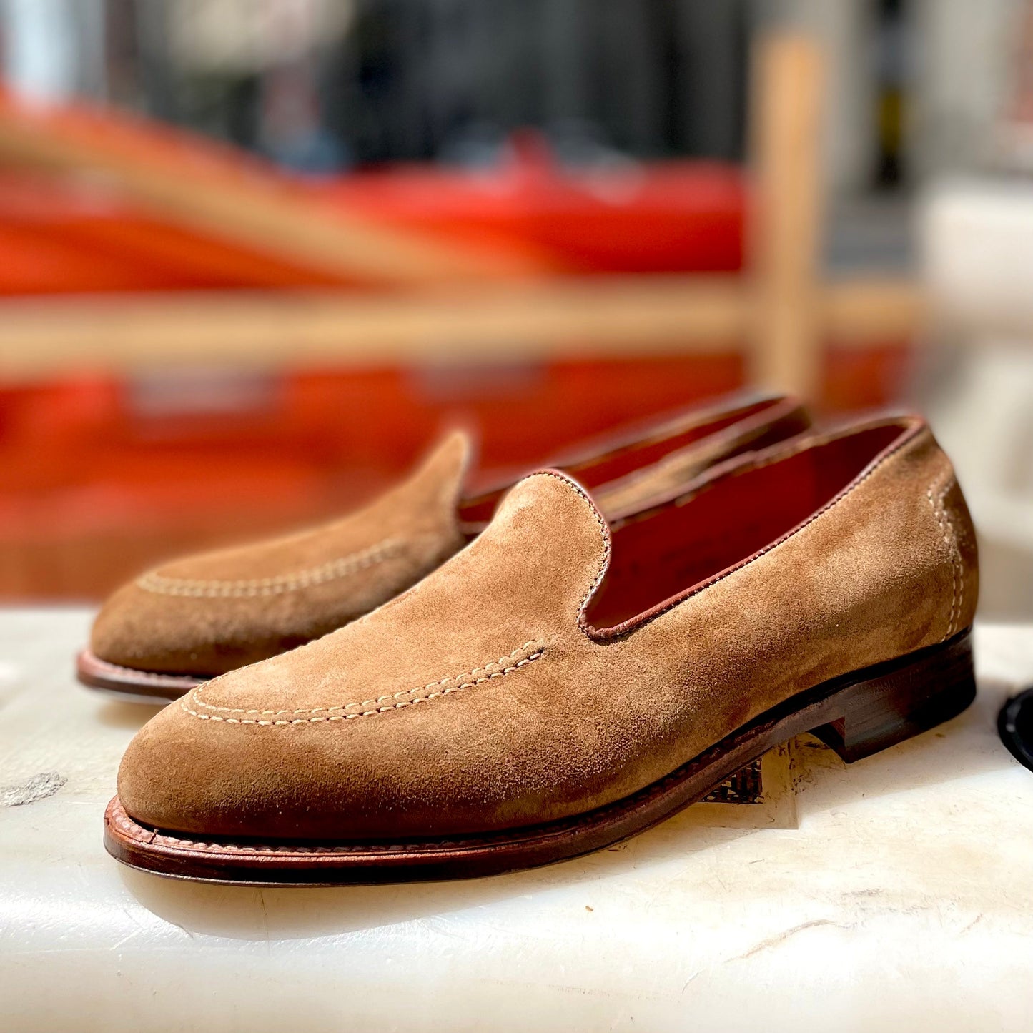 The Alden Madison D2101 - Vanderbilt Slip On in snuff suede, featuring a wooden heel, is showcased on a white surface. The background is slightly blurred with hints of orange and dark colors.