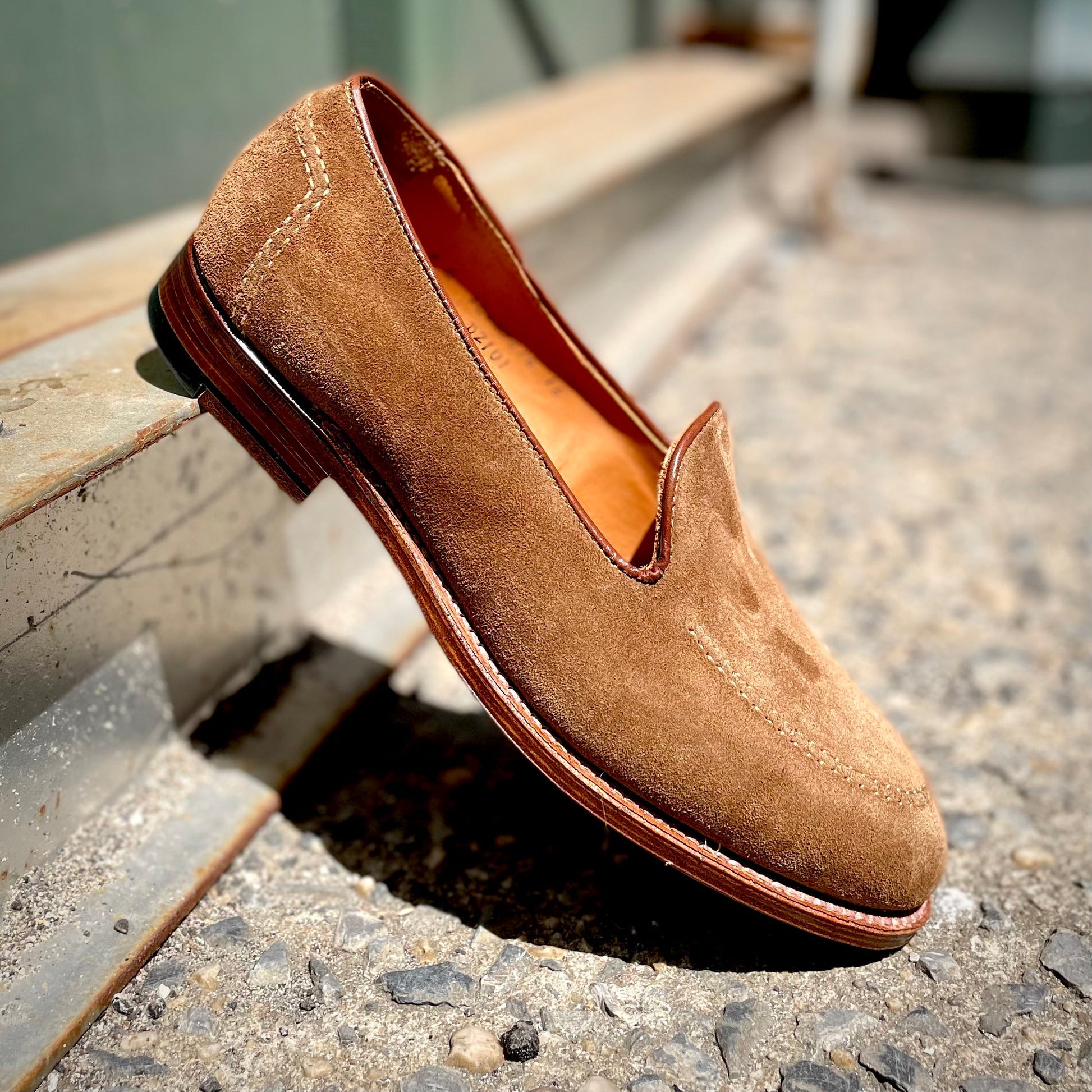 The Alden Madison D2101 - Vanderbilt Slip On in Snuff Suede, featuring a Brooks heel and leather sole, rests angled on a textured concrete surface. Direct sunlight accentuates the visible stitching and rich suede texture.