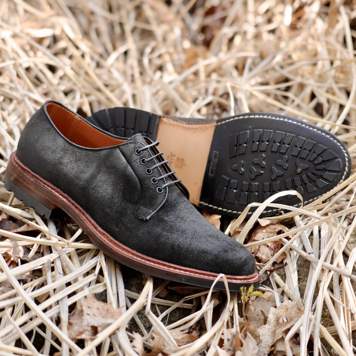 A pair of dark gray Alden Madison D1426C Plain Toe Bluchers in Earth Reverse Chamois rests on dry brown grass. One shoe stands upright showcasing laces, while the other lies sideways revealing its detailed Commando Sole and Barrie Last craftsmanship.