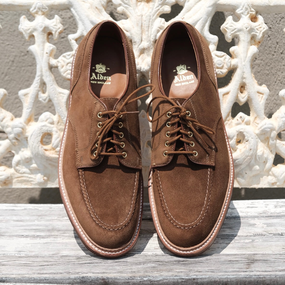 The Alden Madison 24082 - The Quickley in Snuff Suede are showcased on a rustic wooden surface, featuring a moc toe design with contrast stitching and gold eyelets, elegantly standing out against an ornate white background.