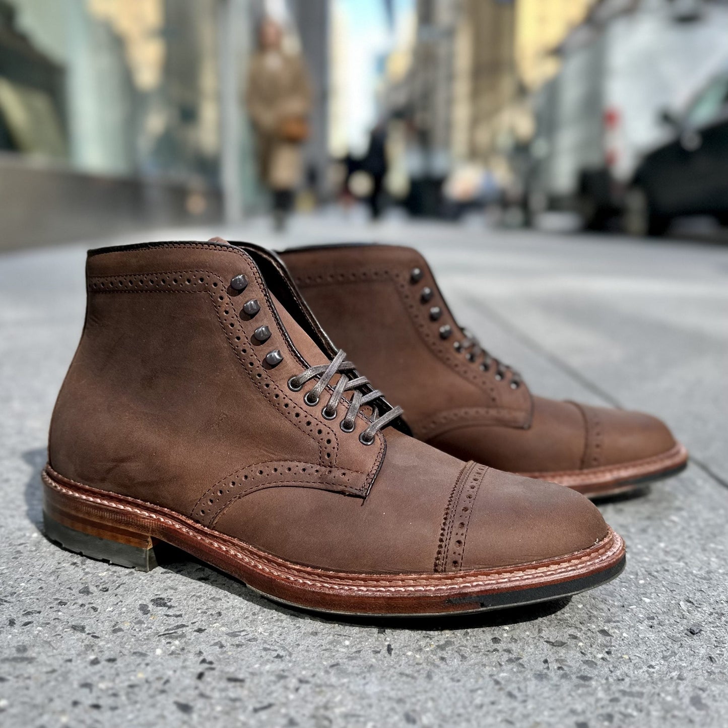 The Alden Madison D2817HC - Straight Tip Shaking Boot in Tobacco Smooth Chamois features detailed stitching and laces on a city sidewalk. The Commando sole ensures durability, with the blurred backdrop highlighting pedestrians and buildings.