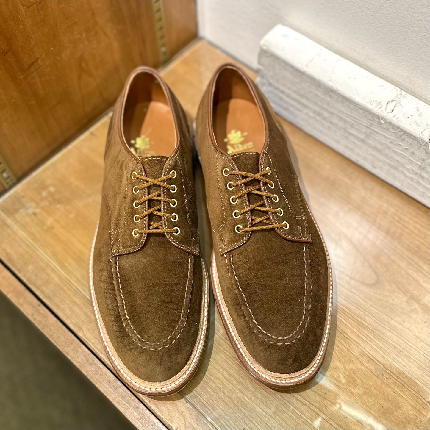 Displayed on a wooden surface against a two-toned wooden and white wall, these Alden Madison 24082 - The Quickley shoes in snuff suede feature tan laces, contrast stitching, and a detailed toe seam.